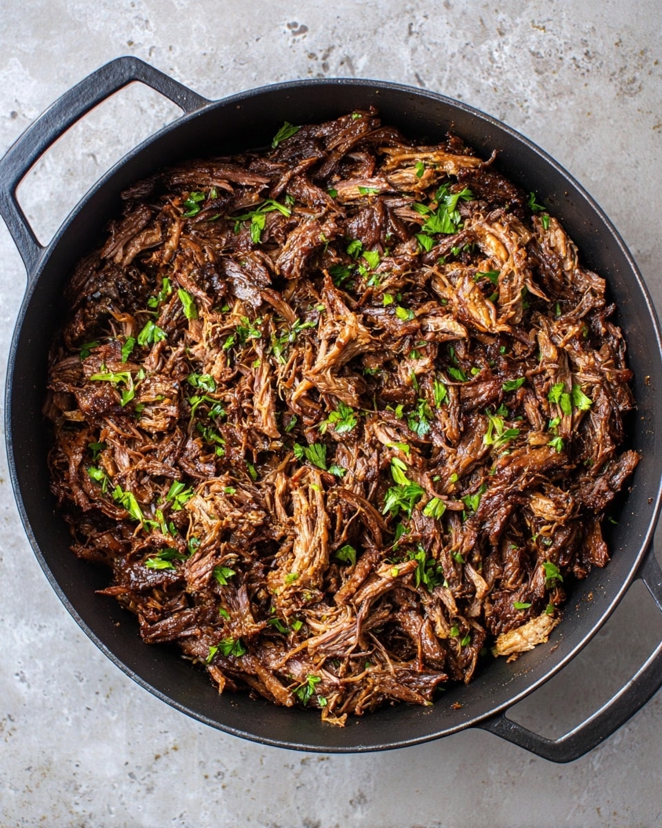 A round black pan filled with shredded dark brown cooked meat with some small bits of light brown fat and green parsley leaves scattered on top, all sitting on a white marbled textured surface. The meat pieces have a moist, shiny texture with varied sizes and some stringy parts visible, showing a rich, savory look. The pan has two handles on opposite sides. Photo taken with an iphone --ar 4:5 --v 7