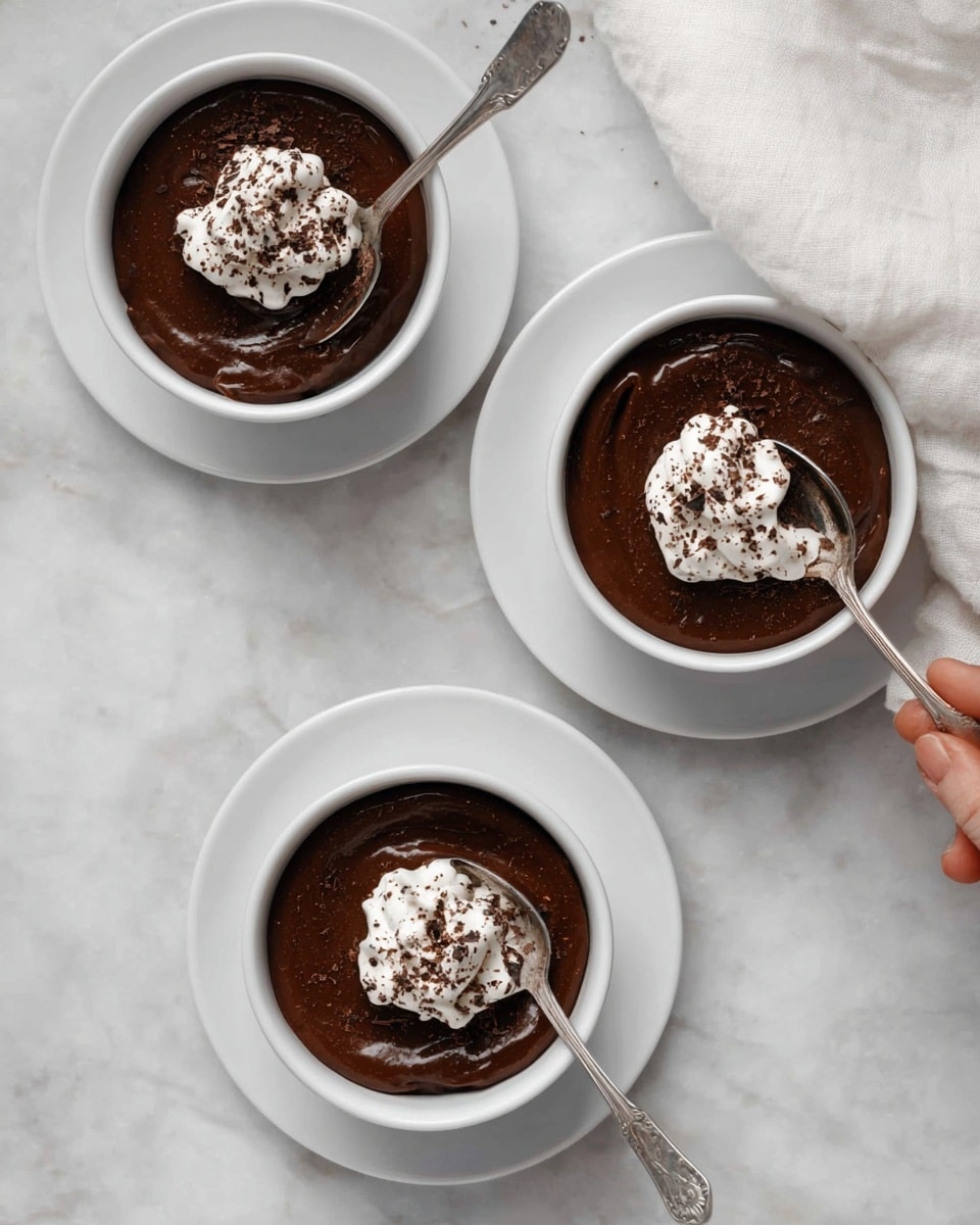 Three white bowls with a smooth dark chocolate pudding inside, each bowl placed on a matching white saucer. On top of each pudding, there is a dollop of white cream sprinkled with dark cocoa powder, with the cream shaped like a flower on one and more spread on others. Three silver spoons rest inside the bowls, one spoon slightly lifted by a woman's hand on the top right bowl. The bowls are arranged on a white marbled surface, and a soft white cloth is partly visible near the top right. photo taken with an iphone --ar 4:5 --v 7