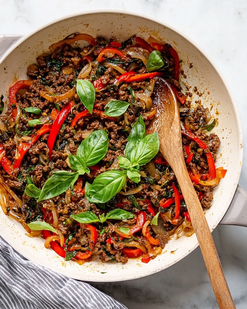 The image shows a white pan filled with cooked ground beef mixed with sliced red bell peppers and onions, all sautéed together creating a colorful mix of brown, red, and translucent white layers. Bright green fresh basil leaves are scattered on top, adding a vibrant contrast. A wooden spoon is resting inside the pan, partially covered in the mixture. The pan is placed on a white marbled surface next to a gray striped cloth. photo taken with an iphone --ar 4:5 --v 7