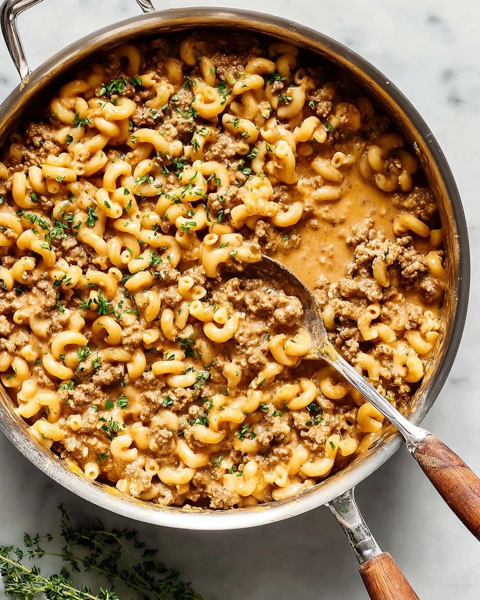 A large silver pan filled with a creamy macaroni dish that includes small pasta tubes mixed with browned ground meat bits throughout. The creamy sauce is light brown and thick, coating the pasta and meat evenly. Small green herb pieces are sprinkled on top, adding contrast and freshness. A silver spoon with a wooden handle rests inside the pan, scooping some of the pasta mixture. The pan is placed on a white marbled surface with a small green herb sprig nearby. photo taken with an iphone --ar 4:5 --v 7