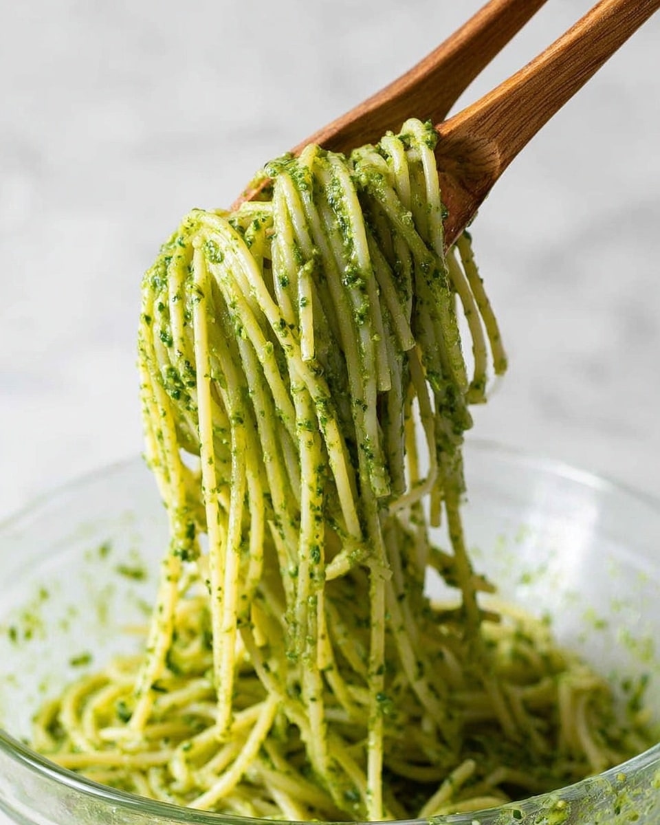 Long thin spaghetti noodles covered with green pesto sauce are lifted by two wooden spoons above a clear bowl. The pesto sauce looks creamy and has small bits of herbs evenly spread on the noodles. The noodles hang down in a thick clump from the spoons, showing a smooth and shiny texture. The background and surface are a white marbled texture. photo taken with an iphone --ar 4:5 --v 7
