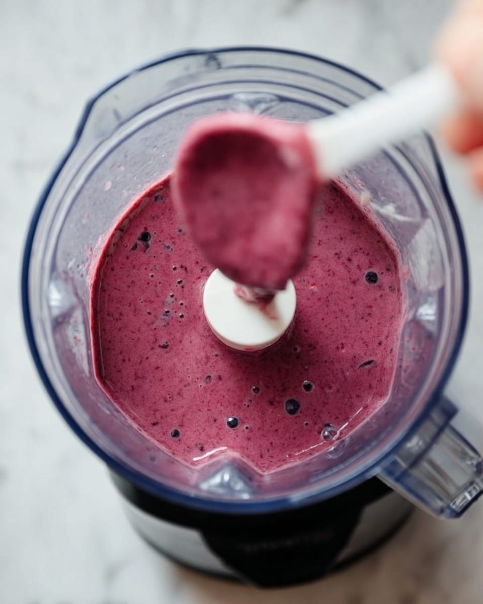 The image shows a close-up top view of a blender filled with thick, purple-red smoothie that has small bubbles and tiny darker specks throughout. The blender is made of clear plastic with a white plastic blade cover inside, and the outside base of the blender is silver with a black bottom edge. The whole blender is placed on a white marbled surface, and a woman's hand is holding a white spoon with some smoothie on it above the blender jar. Photo taken with an iphone --ar 4:5 --v 7