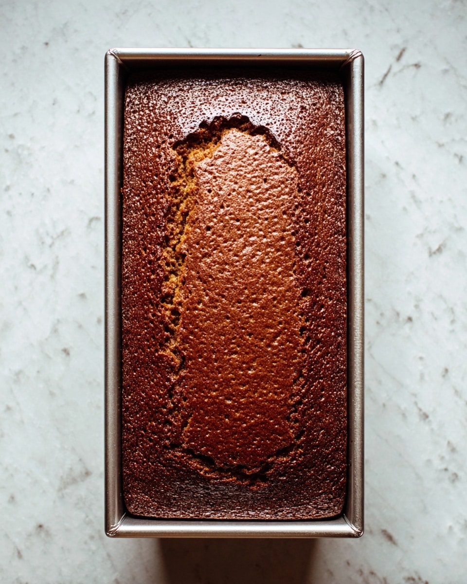 A freshly baked rectangular chocolate cake with a rich brown, slightly cracked top surface sits inside a silver metal loaf pan. The cake's texture looks soft and dense with small air bubbles visible, and the edges are darker and slightly raised compared to the middle. The pan rests on a white marbled surface with subtle grey veins. photo taken with an iphone --ar 4:5 --v 7