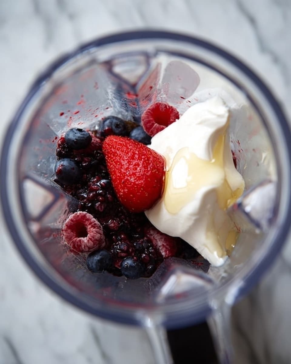 Inside a clear blender jar, there is a layer of mixed dark purple and red berries at the bottom, including blueberries, raspberries, and a single large red strawberry sitting on top. Above the berries, there is a dollop of thick white yogurt with honey drizzled over it, creating a smooth texture. The blender is placed on a white marbled surface. photo taken with an iphone --ar 4:5 --v 7