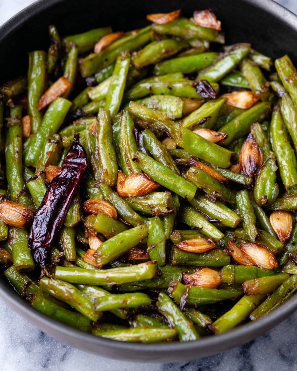 A close-up view of a black pan filled with sautéed green beans and browned garlic slices. The green beans are cut into short pieces and show a mix of light and dark green colors with some parts caramelized to a golden brown. The garlic slices are scattered throughout, showing a toasted light brown color with some darker spots. A whole dark red dried chili pepper is also visible among the green beans. The cooking oils create a shiny, slightly oily texture on the vegetables. The scene is set on a white marbled surface. photo taken with an iphone --ar 4:5 --v 7