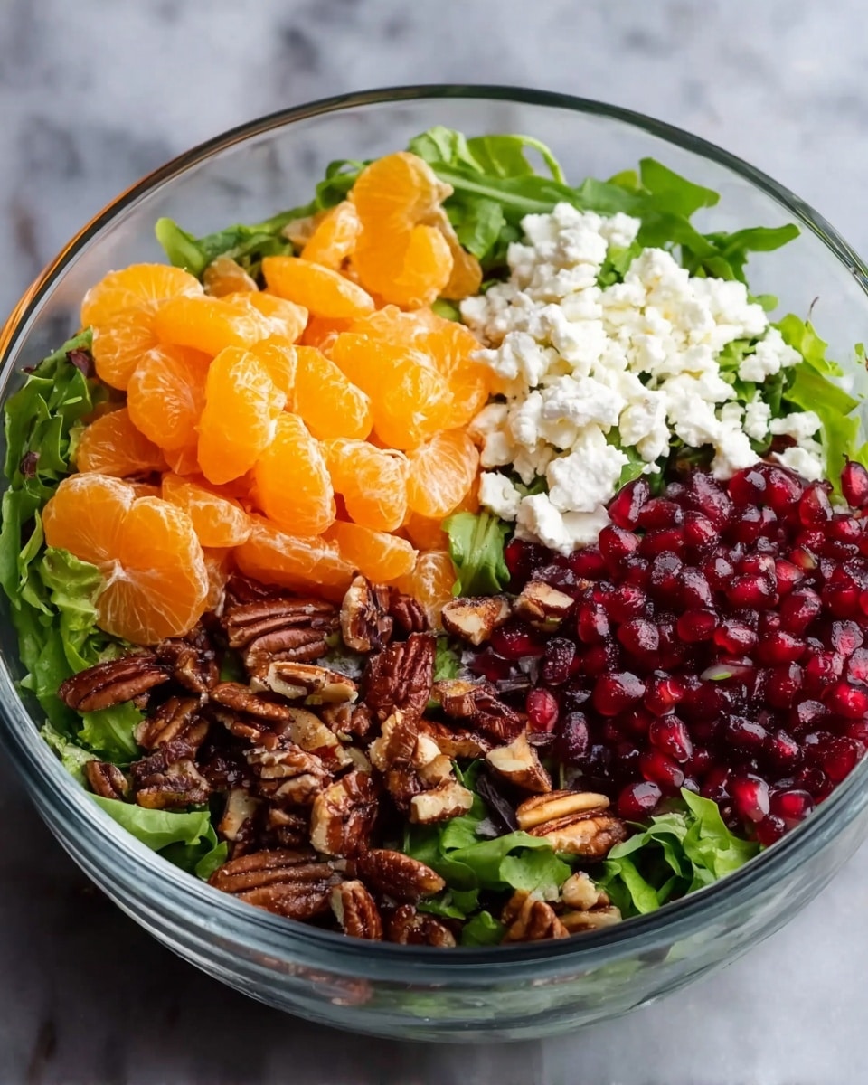 A clear glass bowl filled with a salad showing four main layers from the side and top view: the bottom layer is made of green leafy mix with different shades of green, followed by bright orange mandarin orange segments clustered in one area, next to a pile of small white crumbly cheese, and a section filled with dark brown chopped pecans and bright red pomegranate seeds. The bowl is set on a white marbled surface, photo taken with an iphone --ar 4:5 --v 7