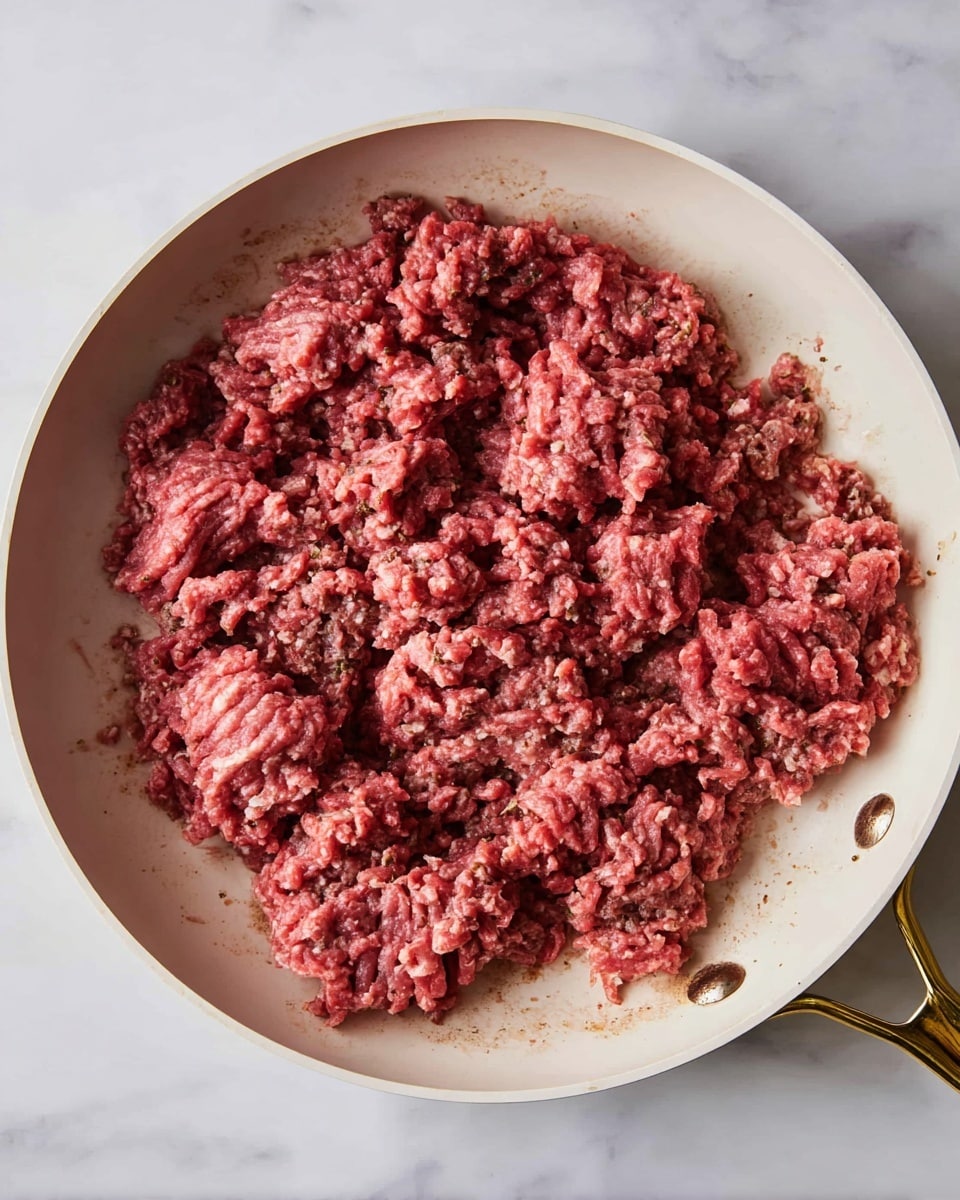 A white pan with a golden handle holds raw ground meat spread unevenly inside. The meat is in one single rough layer, showing a mix of dark red and pink colors with some parts appearing slightly brown. The texture looks crumbly and soft, with small chunks clumped in different sizes across the pan. The pan sits on a white marbled surface. photo taken with an iphone --ar 4:5 --v 7