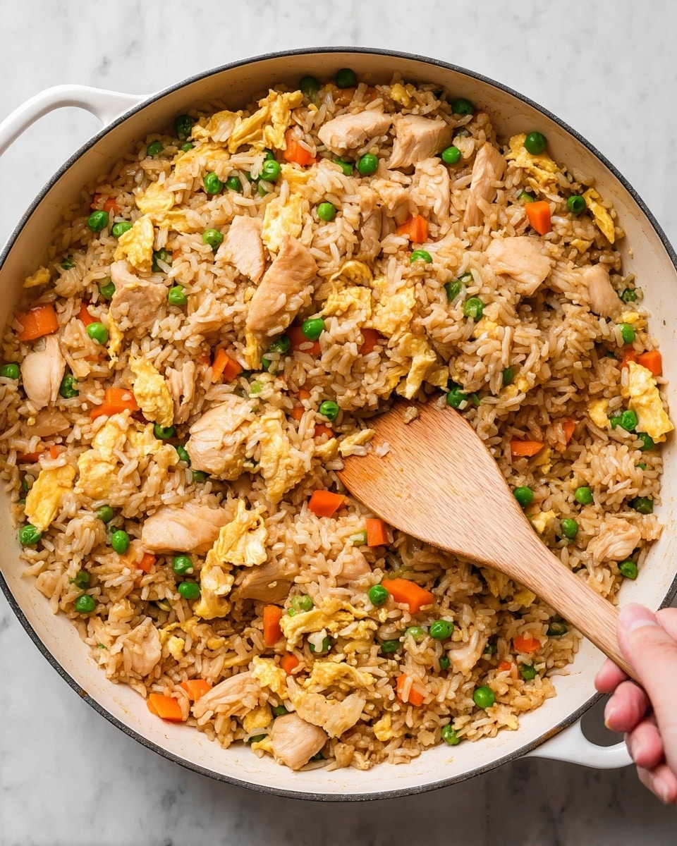 A white pan filled with fried rice showing layers of light brown cooked rice mixed with small green peas, bright orange carrot cubes, and light yellow egg strips spread evenly throughout. Pieces of light brown cooked chicken are scattered within the rice, giving it texture and color contrast. A woman's hand holds a wooden spatula stirring the mixture near the right side of the pan. The pan rests on a white marbled surface photo taken with an iphone --ar 4:5 --v 7