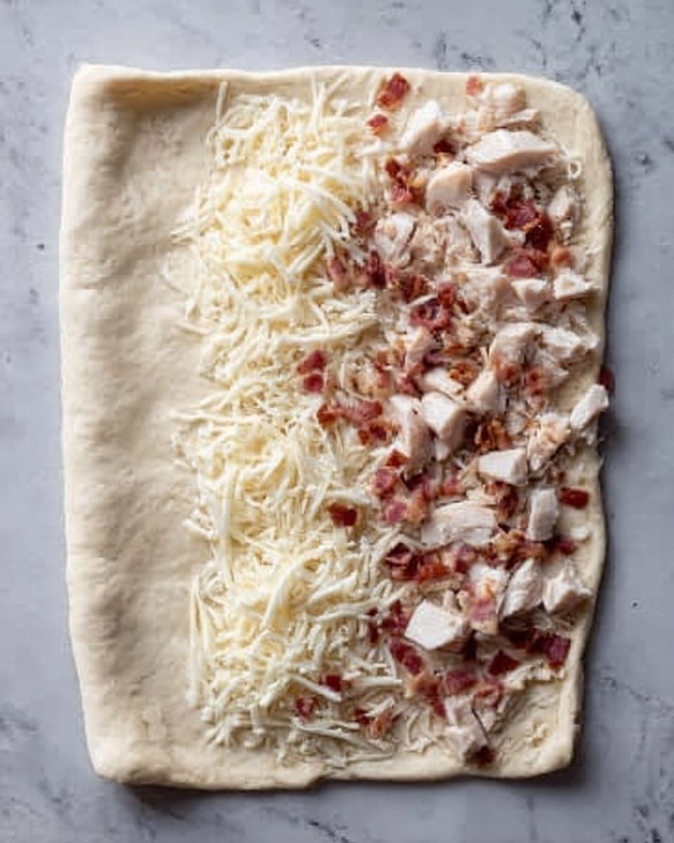 The image shows a rectangular piece of dough laid out on a white marbled surface. On the right side of the dough, there is one layer of small white cubes of cooked chicken, topped by a layer of shredded mozzarella cheese and small pieces of bacon. The left edge of the dough is folded over partially, creating a thick border. The dough looks soft and slightly puffy with a pale cream color. Photo taken with an iphone --ar 4:5 --v 7