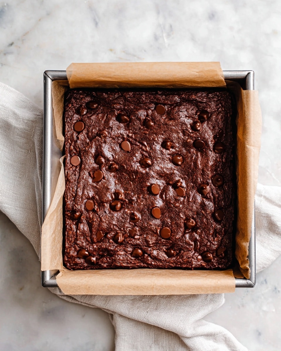 A square metal baking pan holds a thick, dark brown brownie layer with a slightly wrinkled texture on top, dotted with many glossy chocolate chips scattered evenly across the surface. The brownie rests on a piece of light brown parchment paper that extends up on two opposite sides of the pan. The pan sits on a white marbled surface with a soft, white cloth partially visible underneath. photo taken with an iphone --ar 4:5 --v 7