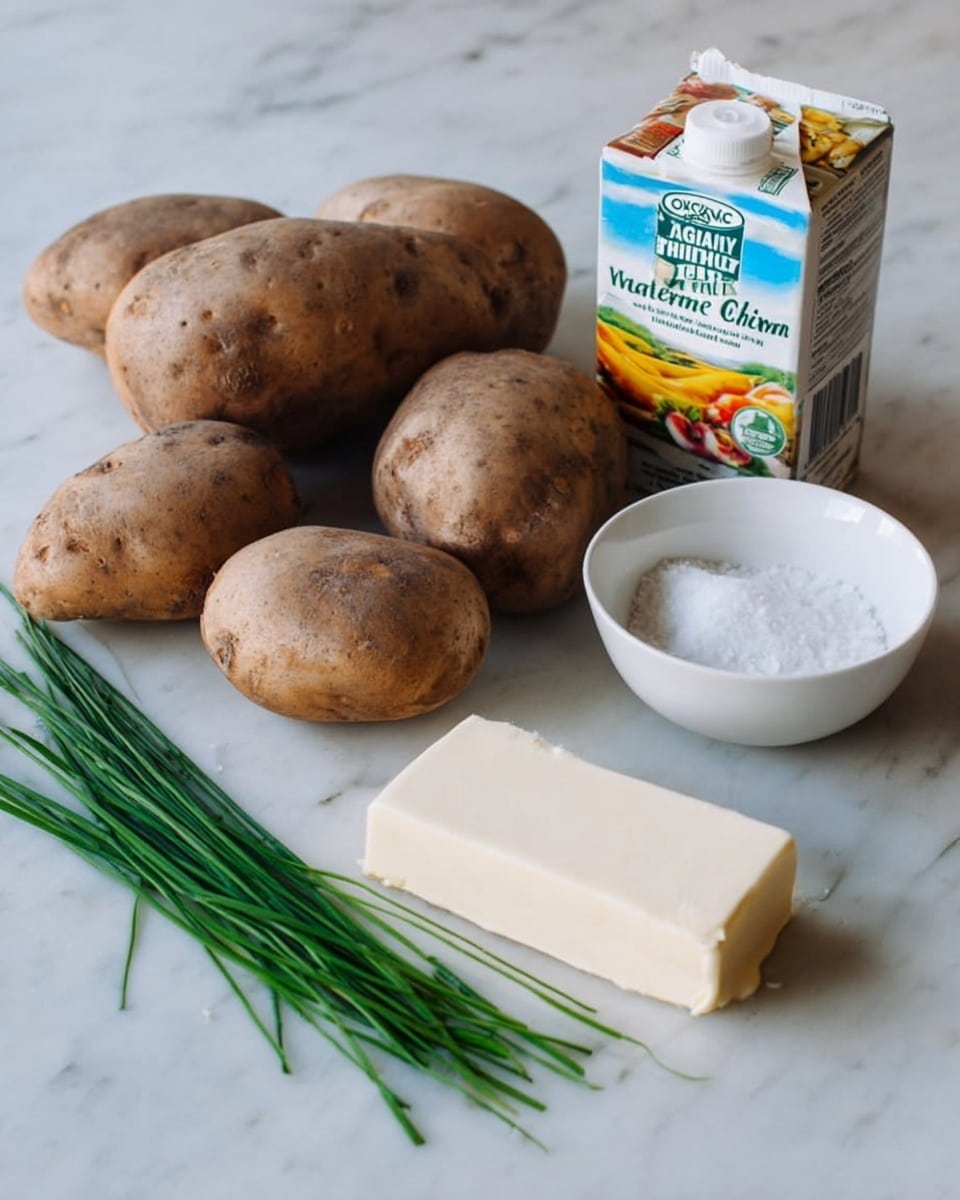 The image shows five whole russet potatoes with rough brown skin placed on a white marbled surface. Below the potatoes, there is a small bunch of green chives laid flat. To the right of the potatoes, there is a white bowl filled with white salt grains. In front of the salt bowl, there is a rectangular stick of unsalted butter wrapped in cream-colored paper with black text. Behind the butter and salt, there is a carton of Organic Valley heavy whipping cream with bright yellow, blue, and white colors with images of cream and fruit on it. photo taken with an iphone --ar 4:5 --v 7