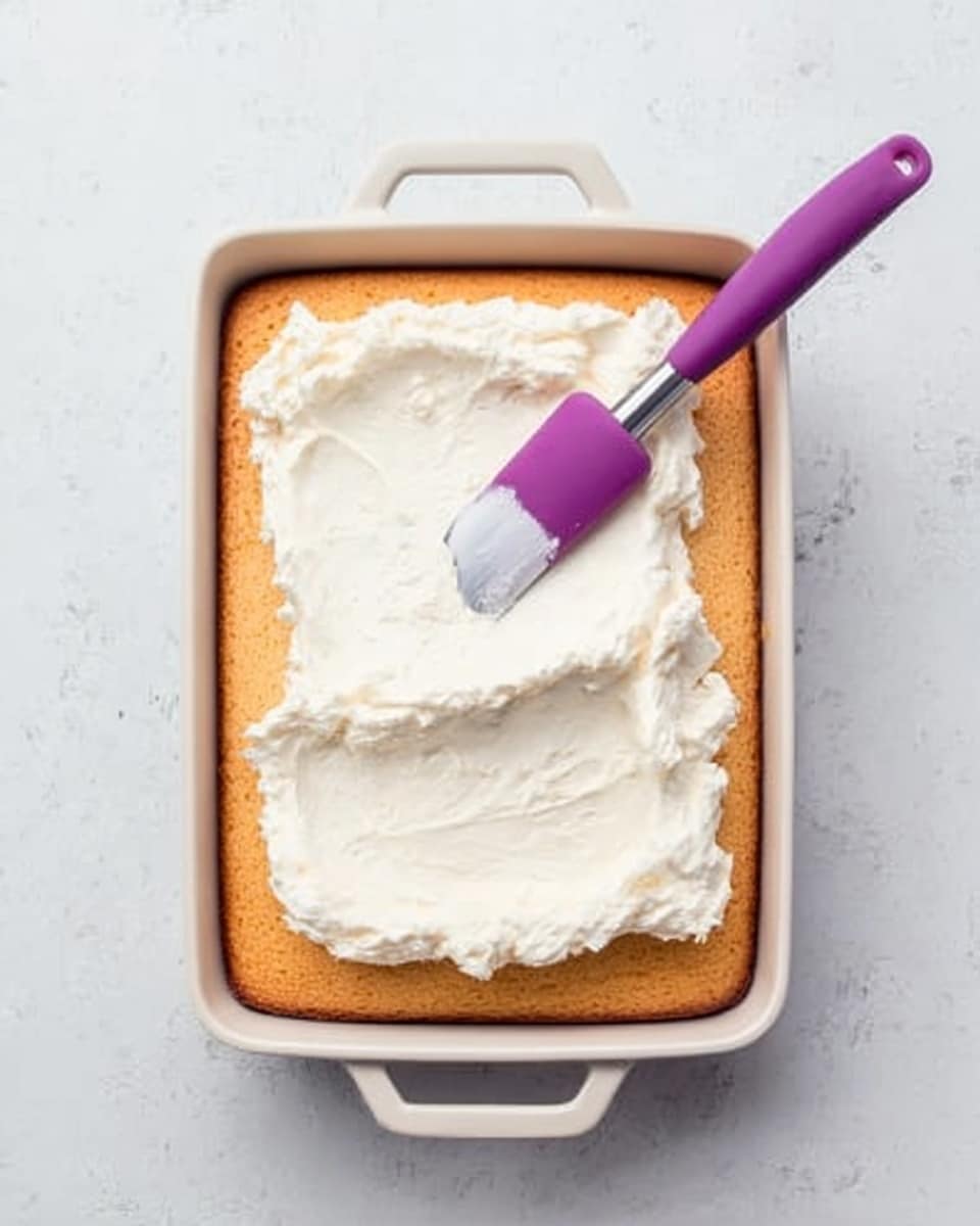 A rectangular white baking dish with handles holds a single layer of golden-brown cake. On top of the cake, there is a thick layer of white, creamy frosting being spread evenly with a purple-handled spatula. The background surface is a white marbled texture, creating a clean, bright setting. Photo taken with an iphone --ar 4:5 --v 7