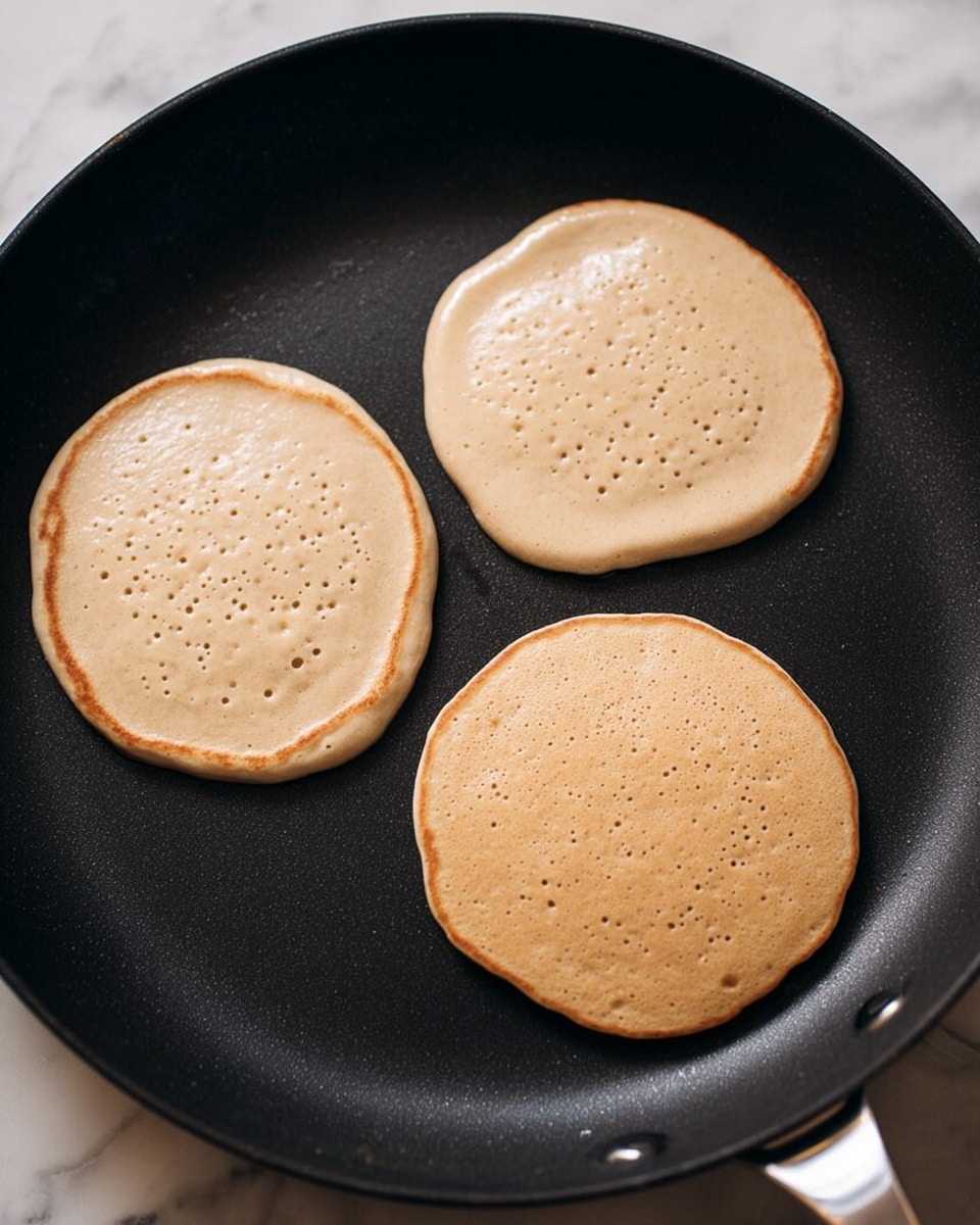 The image shows three light brown pancake batter circles cooking on a black pan. The pancakes are evenly spaced, with small bubbles forming on their surfaces, giving them a slightly textured look. The pan is smooth and flat, with a silver handle partially visible on the right side. The background surface has been changed mentally to a white marbled texture. photo taken with an iphone --ar 4:5 --v 7