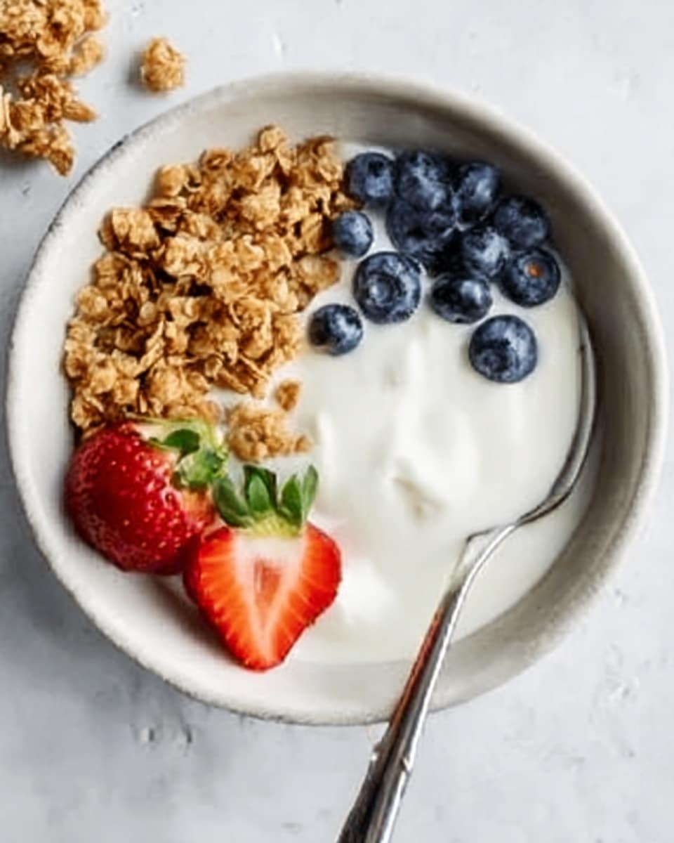A white bowl on a white marbled surface holds a creamy, smooth yogurt base filling most of the bowl space. On the left side, there is a cluster of golden-brown granola with a crunchy texture. On the right, there is a group of fresh blueberries with a dark blue and slightly shiny surface. In the front, two whole strawberries with a bright red color and green leaves sit partially on the yogurt. A silver spoon with a long handle rests inside the bowl, leaning against the edge, with a woman's hand about to hold it. Photo taken with an iphone --ar 4:5 --v 7