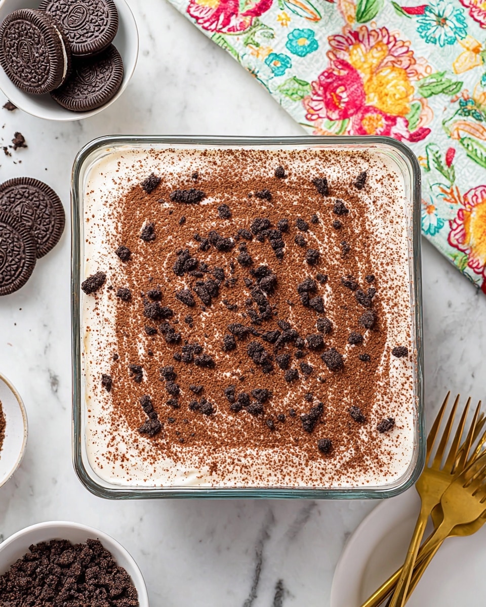 A square glass dish holds a dessert with two visible layers: the bottom layer is creamy white, smooth and thick, while the top layer is light brown cocoa powder evenly dusted, sprinkled with small, dark chocolate cookie crumbles scattered across the surface. The dish is placed on a white marbled surface with a few whole chocolate sandwich cookies in the upper left corner, a colorful floral cloth in the top right corner, a small white bowl with more crumbled cookies in the bottom left, and a white plate with golden forks at the bottom right. The overall look is neat and inviting. photo taken with an iphone --ar 4:5 --v 7