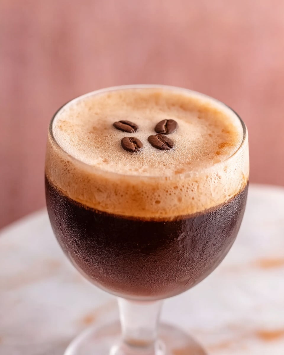 A close-up of a clear glass cup filled with dark brown coffee at the bottom, topped with a thick, light tan foam layer covering the entire surface. Three medium brown whole coffee beans are placed on top of the foam in the center. The cup is set on a white marbled surface with a soft pinkish background blurred out. Photo taken with an iphone --ar 4:5 --v 7