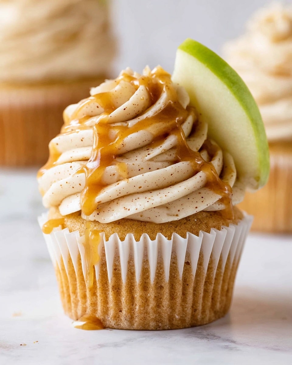 A golden brown cupcake in a white paper wrapper sits on a white marbled surface, topped with a tall swirl of light beige frosting speckled with tiny darker dots, creating a creamy texture. Drizzled over the frosting is a shiny, amber-colored sauce that flows softly down the swirls and over the cupcake edge. A thin slice of green apple with a pale yellow center is placed vertically, slightly tucked into the frosting at the back. In the blurred background, another similar cupcake is partially visible. Photo taken with an iphone --ar 4:5 --v 7