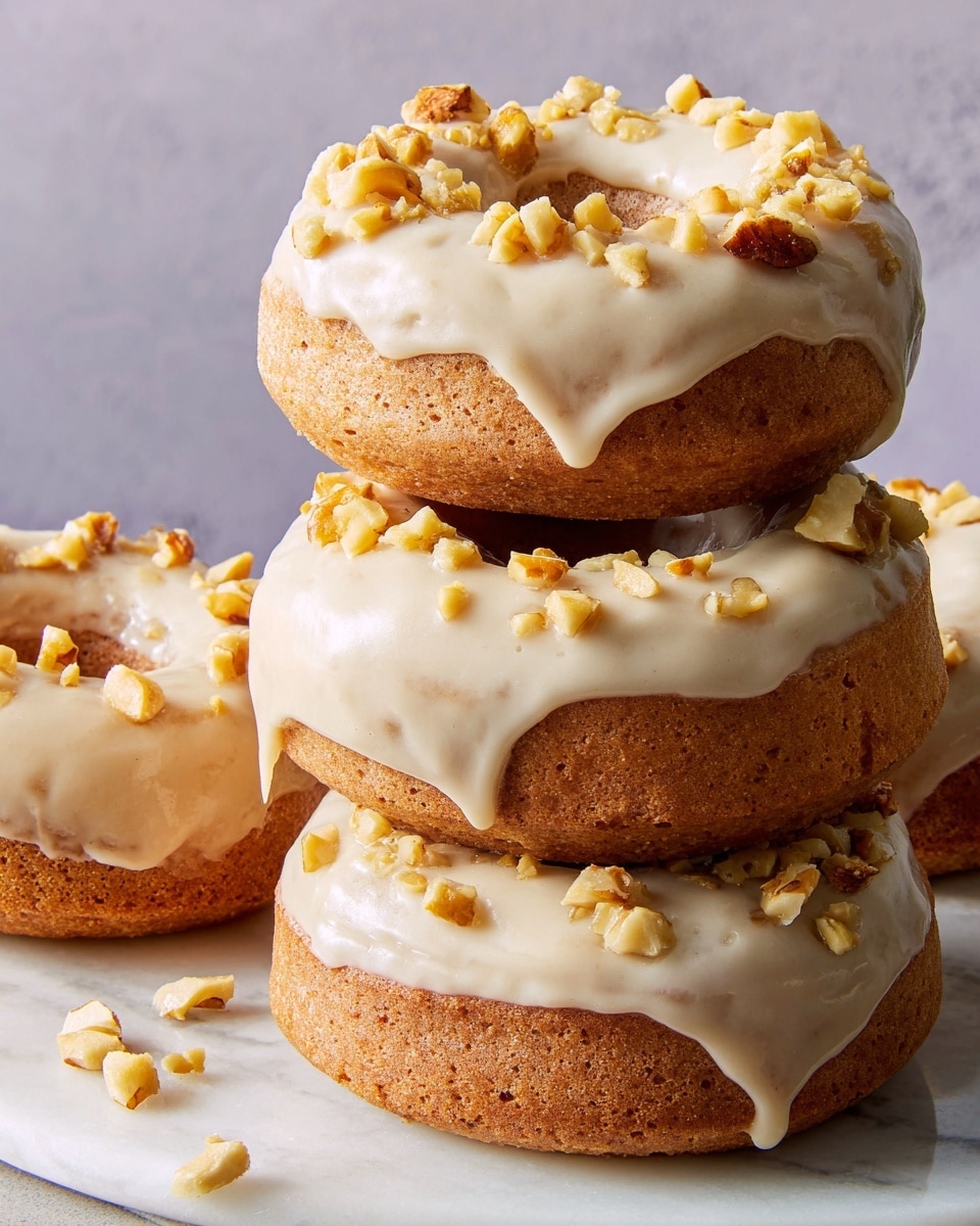 A close-up of four light brown donuts stacked on a white marbled surface with a plain gray wall behind. Each donut has a smooth layer of off-white icing on top that drips slightly down the sides. Small pieces of chopped nuts with yellowish-brown tones are sprinkled over the icing, adding texture and contrast. The donuts look soft and moist with a slightly rough surface underneath the icing. Photo taken with an iphone --ar 4:5 --v 7