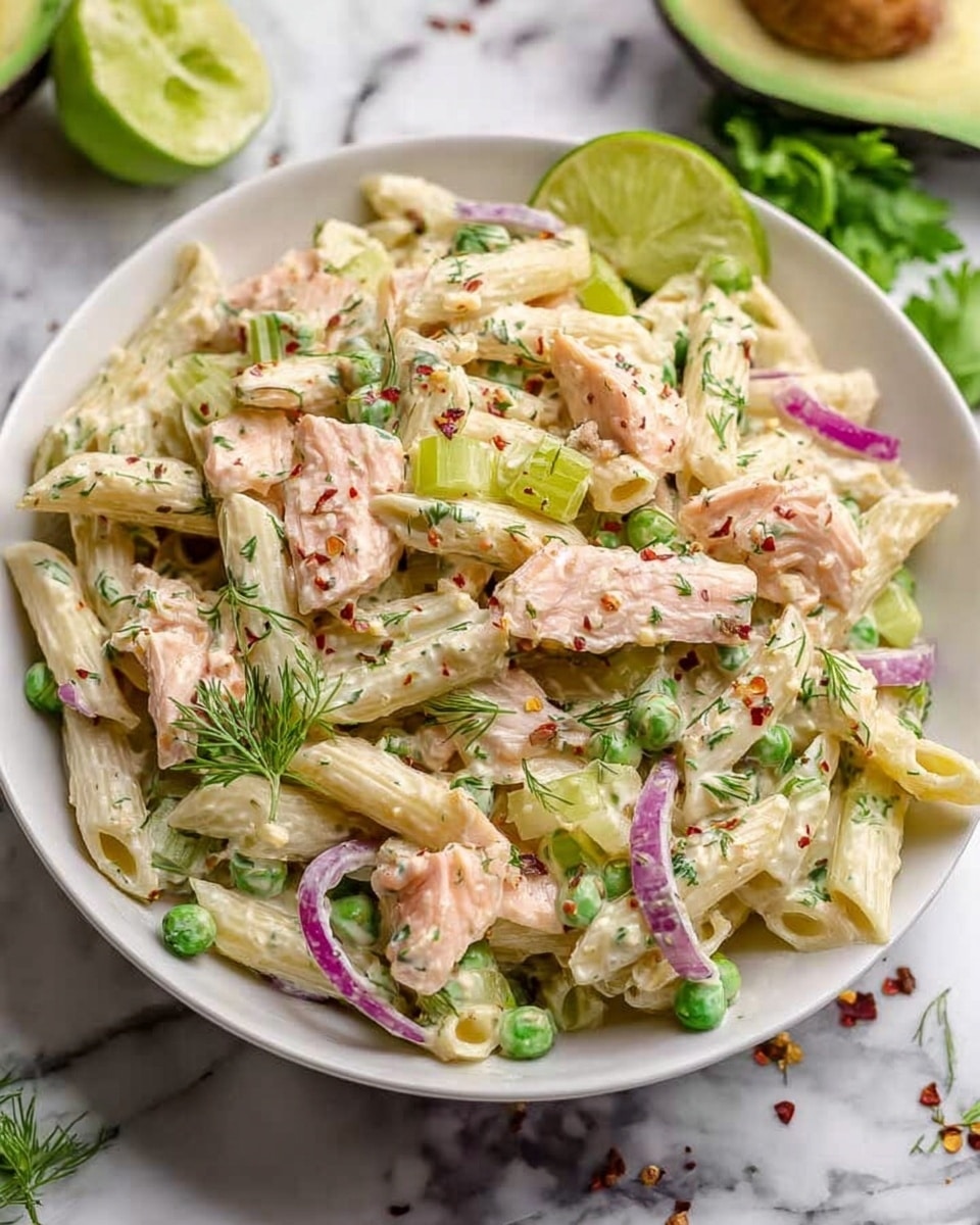 A white bowl filled with a creamy pasta salad layered with penne pasta coated in a light white sauce, soft light pink pieces of salmon, thin slices of purple onion, and small green peas scattered throughout. There are pale green celery slices mixed in, and the dish is sprinkled with finely chopped fresh dill and small red chili flakes for color. A lime slice is placed on the side of the bowl. The bowl sits on a white marbled surface with green celery leaves and half an avocado visible in the background. photo taken with an iphone --ar 4:5 --v 7