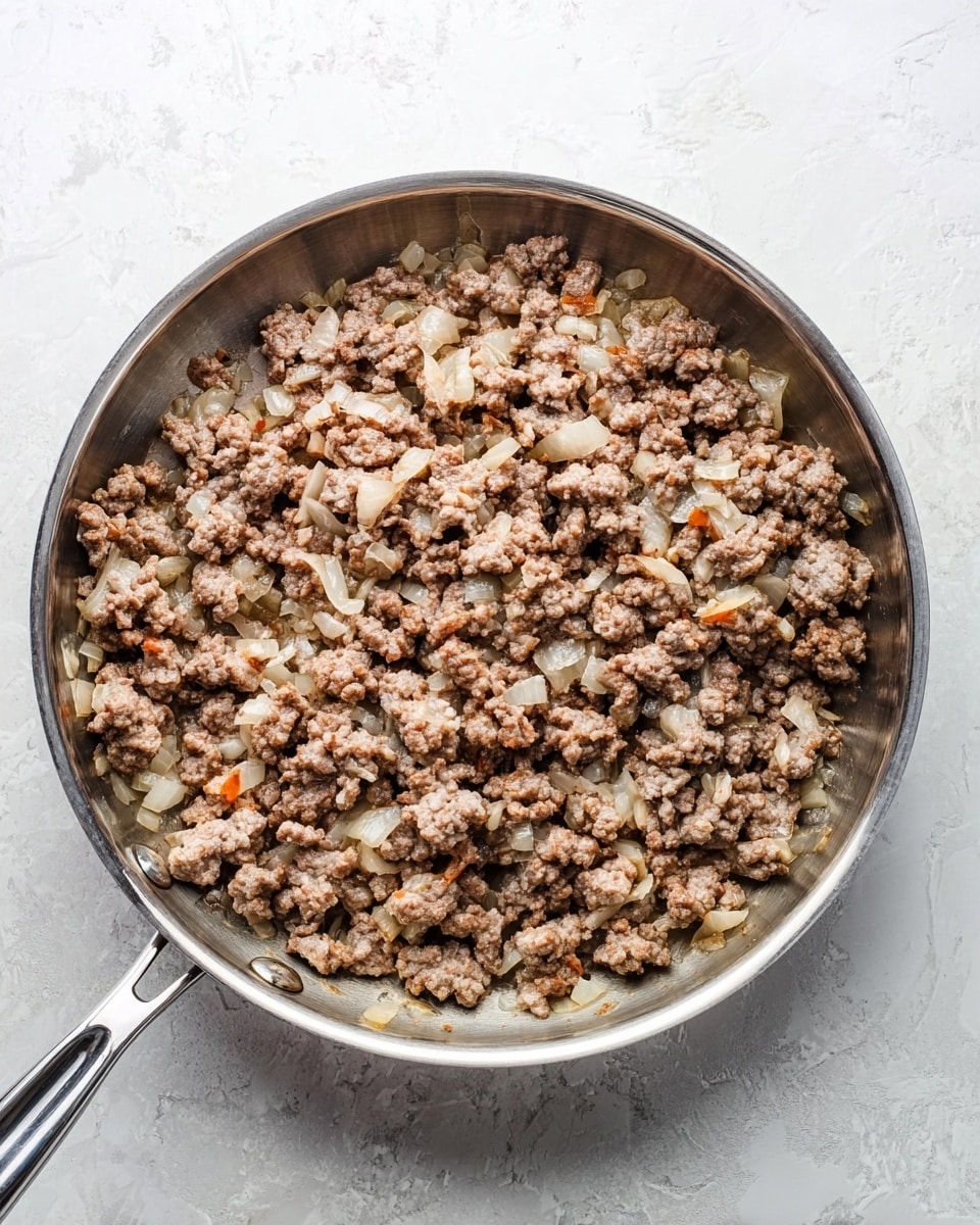 A silver metal frying pan filled with cooked ground meat and chopped onions. The mixture is spread evenly inside the pan, showing light brown chunks of meat mixed with translucent and slightly browned small onion pieces. The pan has two handles, one long and one short. The background is a white marbled texture. Photo taken with an iphone --ar 4:5 --v 7