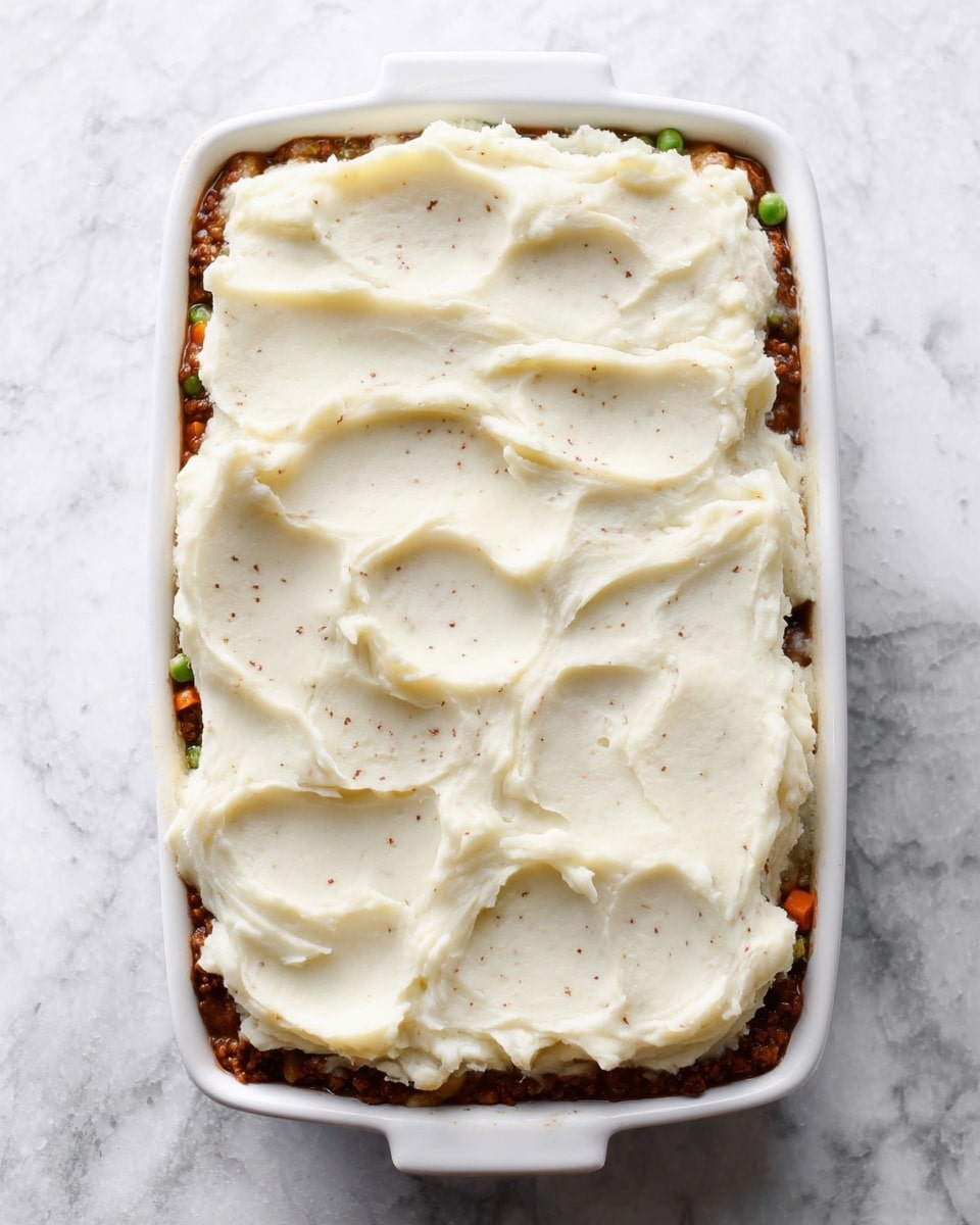 A white rectangular ceramic dish filled with two visible layers, the bottom layer is a brown, meaty mixture with small bits of vegetables peeking through the edges, and the top layer is smooth, thick mashed potatoes spread unevenly with visible swirls and small brown specks, covering the entire surface. The dish is placed on a white marbled surface. photo taken with an iphone --ar 4:5 --v 7