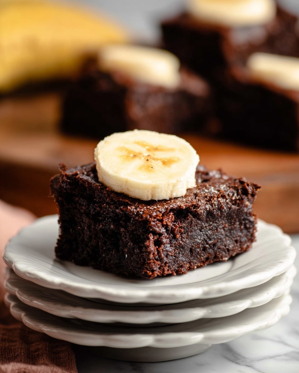 A single square piece of dark brown chocolate brownie sits on top of a stack of five white scalloped plates. The brownie has a textured, slightly cracked surface with a matte finish. On top of the brownie, there is a thick, round slice of pale yellow banana with light brown specks in the center. In the background, more pieces of similar dark brownies can be seen on a wooden surface, blurred out to keep focus on the front piece. The whole scene is set on a white marbled texture. photo taken with an iphone --ar 4:5 --v 7