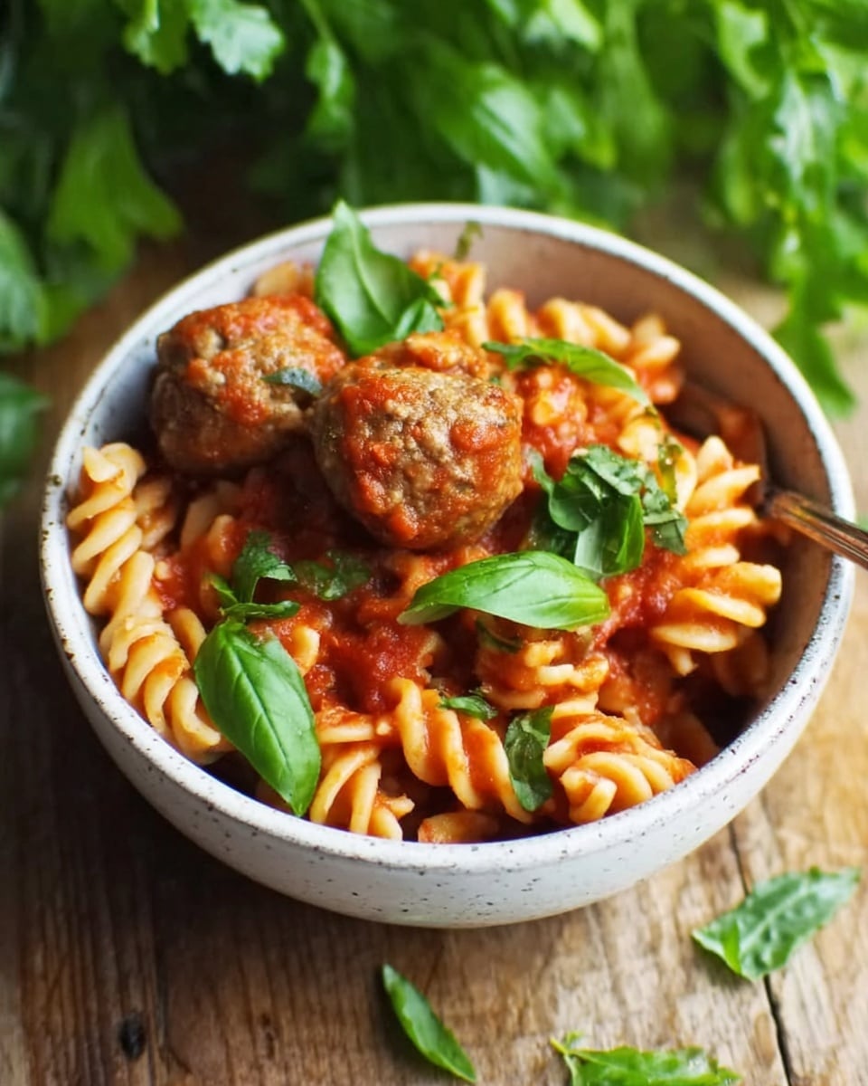 A white ceramic bowl filled with spiral pasta mixed with a thick red tomato sauce, topped with three brown meatballs. Fresh green basil leaves are placed on top and scattered around the bowl. The bowl is sitting on a wooden surface, with blurred green leaves in the background. Photo taken with an iphone --ar 4:5 --v 7