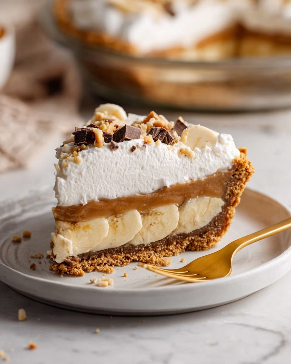 A slice of pie sits on a white plate with a golden fork beside it. The pie has four layers: a light brown crumbly crust at the bottom, a thick caramel-colored filling above it, a layer of sliced bananas in the middle, and a tall layer of white whipped cream on top. The whipped cream is sprinkled with small crunchy brown and tan bits. In the background, part of the remaining pie is visible on a white marbled surface. Photo taken with an iphone --ar 4:5 --v 7