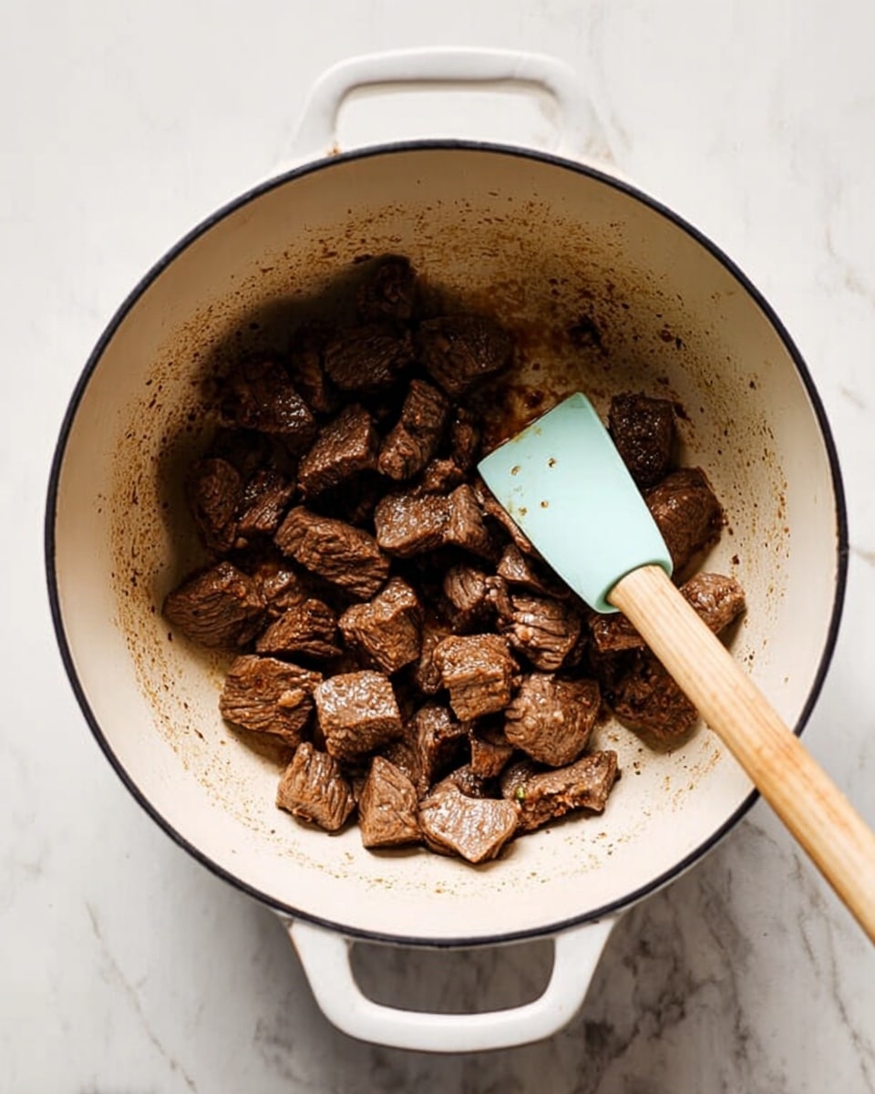 Inside a large white enamel pot with light brown stains around the edges, there are many small, irregular chunks of browned meat clustered at the center bottom. A spatula with a light blue silicone head and a wooden handle is partially under some meat pieces, angled to the right. The pot sits on a white marbled surface. photo taken with an iphone --ar 4:5 --v 7