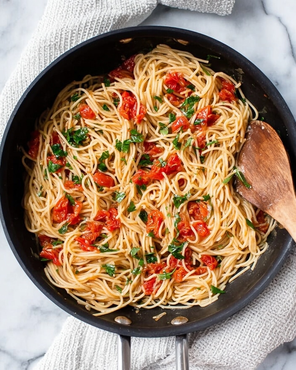A black pan filled with cooked spaghetti pasta mixed with small pieces of cooked red tomatoes and chopped green herbs spread evenly on top, a wooden spoon rests on the right side of the pan near the edge. The pan is placed on a white marbled surface, next to a white cloth with a subtle textured pattern. The colors are natural with the light brown of the pasta, bright red of the tomatoes, and fresh green of the herbs, showing a simple and fresh pasta dish photo taken with an iphone --ar 4:5 --v 7