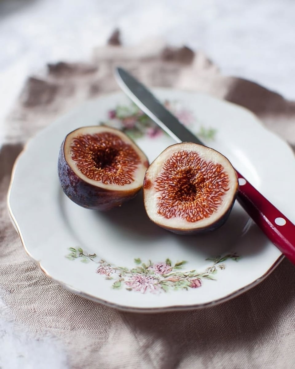 Two halves of a fig with a deep purple outer skin and a textured, reddish-brown inside lie on a white plate with delicate floral designs. The fig halves are positioned side by side in the center of the plate. Behind them rests a small knife with a silver blade and a red handle. The plate sits on a light fabric cloth with a slightly rough texture, placed over a white marbled surface. photo taken with an iphone --ar 4:5 --v 7