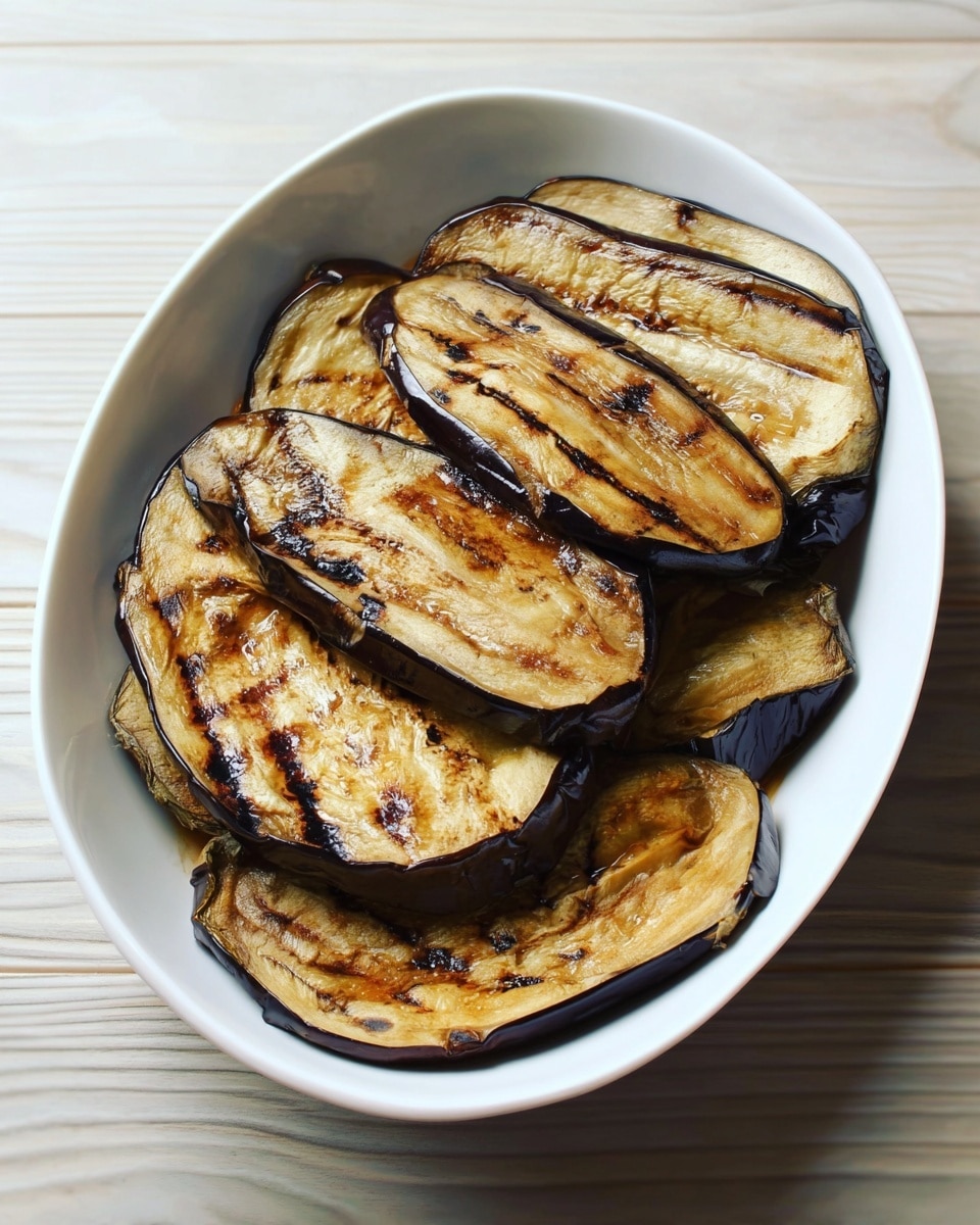 A white oval bowl filled with several large, grilled eggplant slices stacked on top of each other. The eggplant slices have a golden brown color with dark grill marks and slightly caramelized edges, showing a soft and tender texture. The bowl sits on a white marbled surface with a light wood pattern visible around the bowl. photo taken with an iphone --ar 4:5 --v 7
