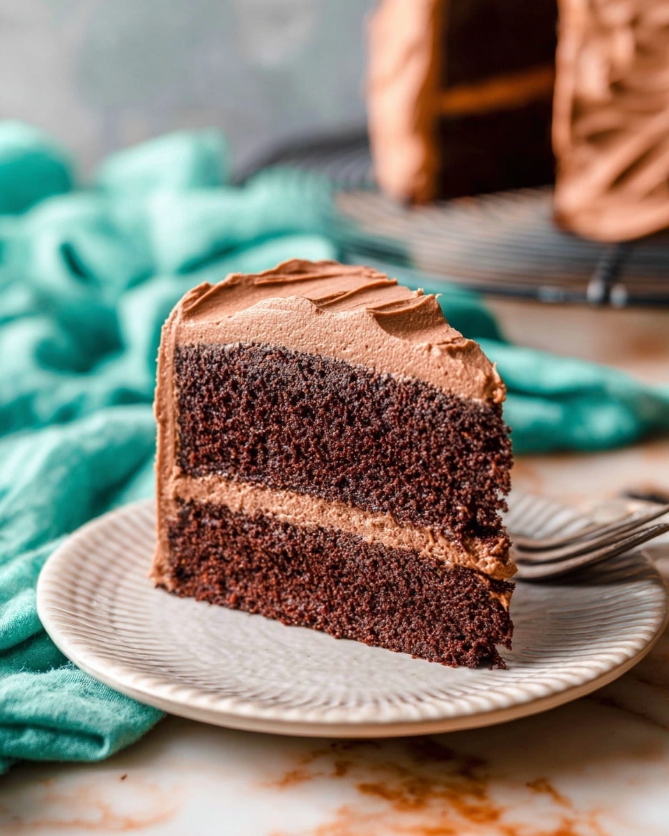 A slice of layered chocolate cake sits on a white plate with a textured edge. The cake has two thick layers of dark brown moist chocolate sponge, separated by a middle layer of smooth, light brown chocolate frosting. The top and sides of the cake are covered in the same creamy chocolate frosting, with visible swirls and soft peaks. In the background, there is a soft focus turquoise cloth and part of the full chocolate cake on a cooling rack. The scene is set on a white marbled surface with warm, natural light. Photo taken with an iphone --ar 4:5 --v 7