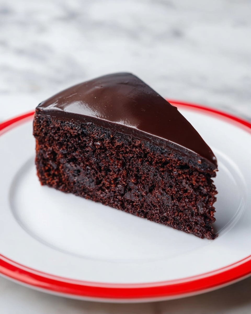 A single slice of dark chocolate cake with one thick layer of moist, crumbly chocolate sponge and a smooth, shiny dark chocolate glaze covering the top and sides, placed on a white plate with a red rim on a white marbled surface. Photo taken with an iphone --ar 4:5 --v 7