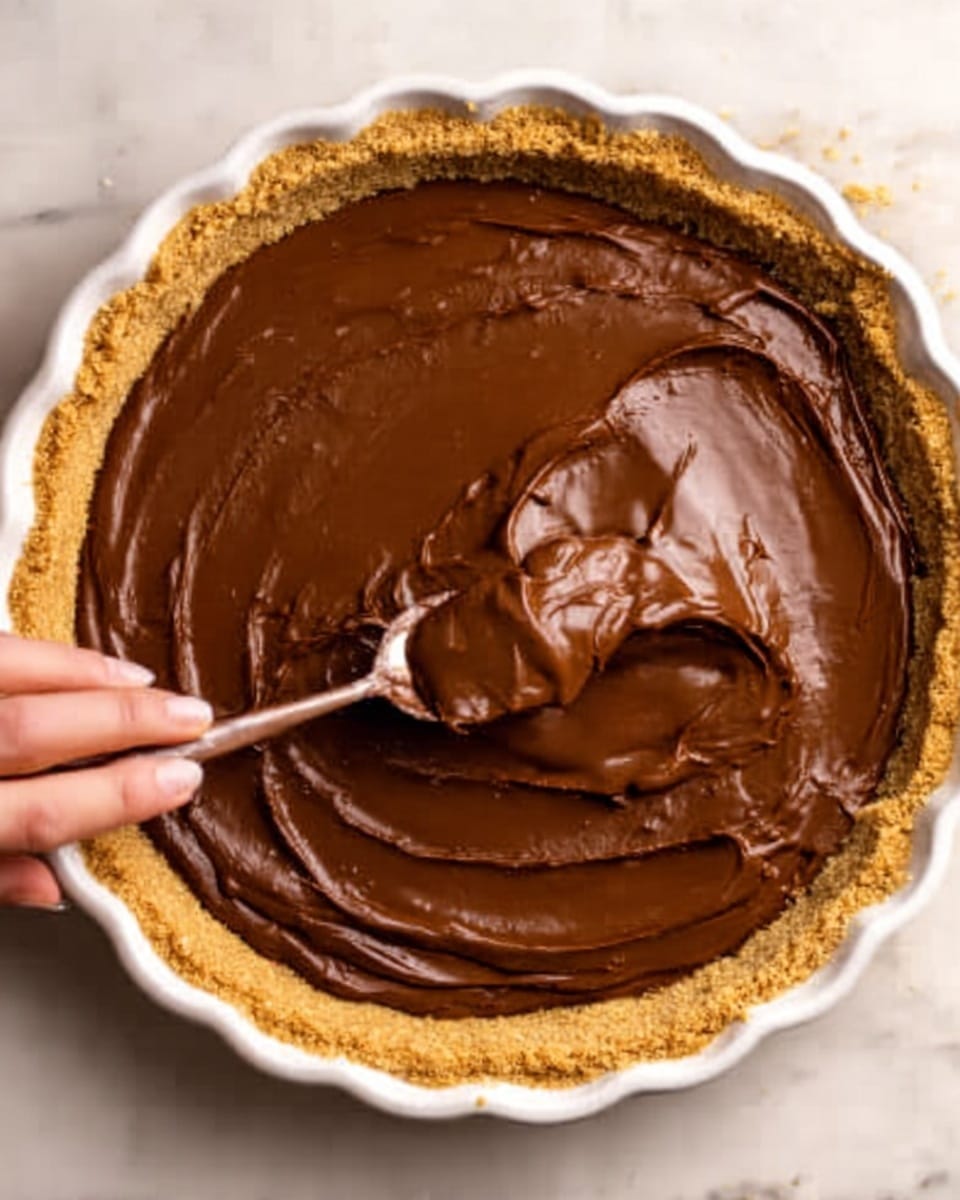 A white tart pan filled with a thick, smooth, shiny chocolate layer that is being spread evenly inside the crust. The crust is light brown, crumbly, and surrounds the chocolate layer from the sides. A woman's hand holding a spoon with chocolate is lifting some of the chocolate from the center, showing the creamy texture. The tart pan is placed on a white marbled surface. photo taken with an iphone --ar 4:5 --v 7