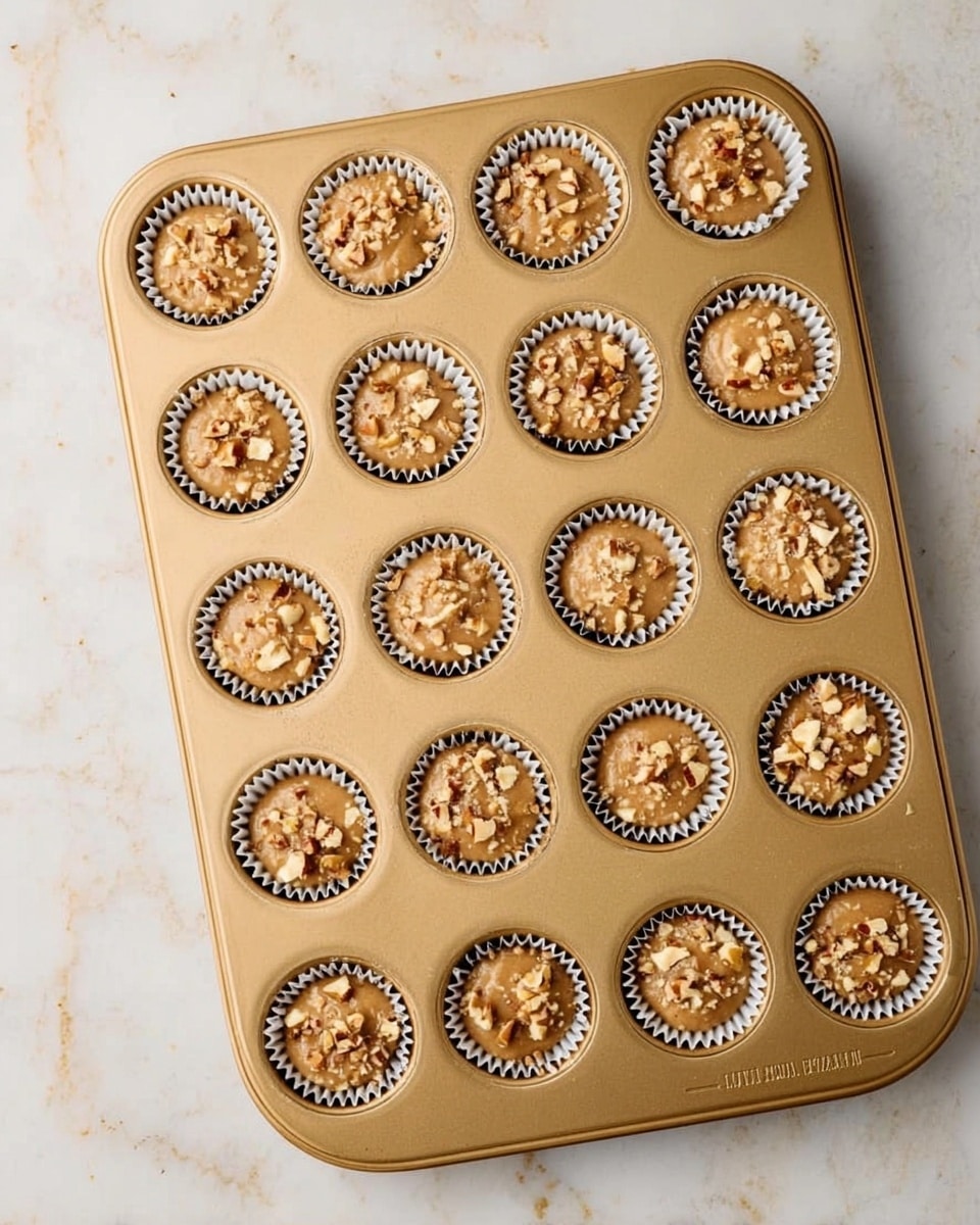 A gold colored muffin tray holds 24 paper cupcake liners filled with light brown muffin batter topped with small pieces of chopped nuts. The batter is unevenly spread in each liner. The tray is set on a white marbled surface. Photo taken with an iphone --ar 4:5 --v 7