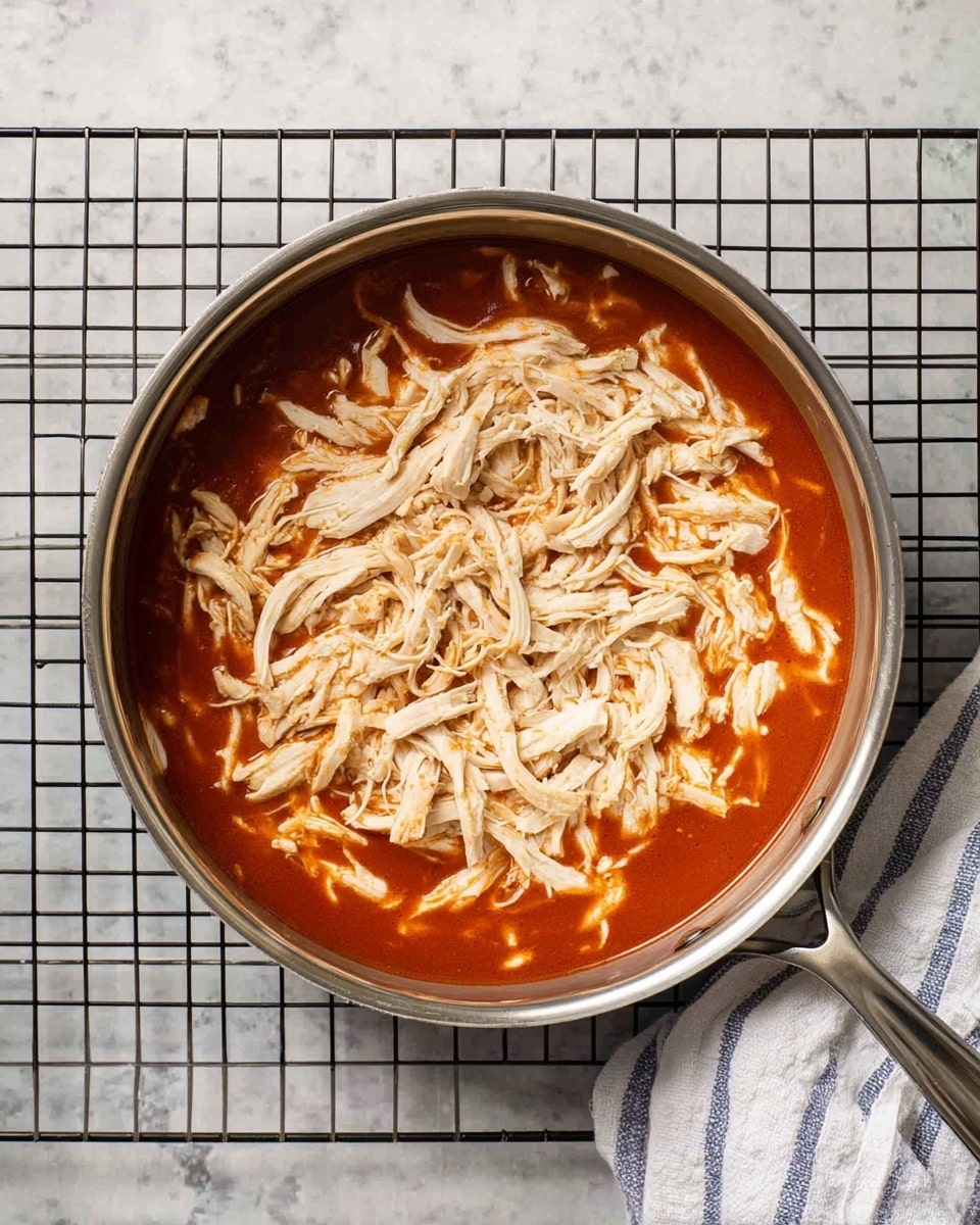 A metal pan with two layers inside is placed on a black cooling rack over a white marbled surface. The bottom layer is a smooth, rich red sauce that fills the pan evenly. On top, there is a thick layer of shredded light beige chicken covering most of the sauce. A white and blue striped cloth is partially visible on the right side. The photo was taken with an iphone --ar 4:5 --v 7