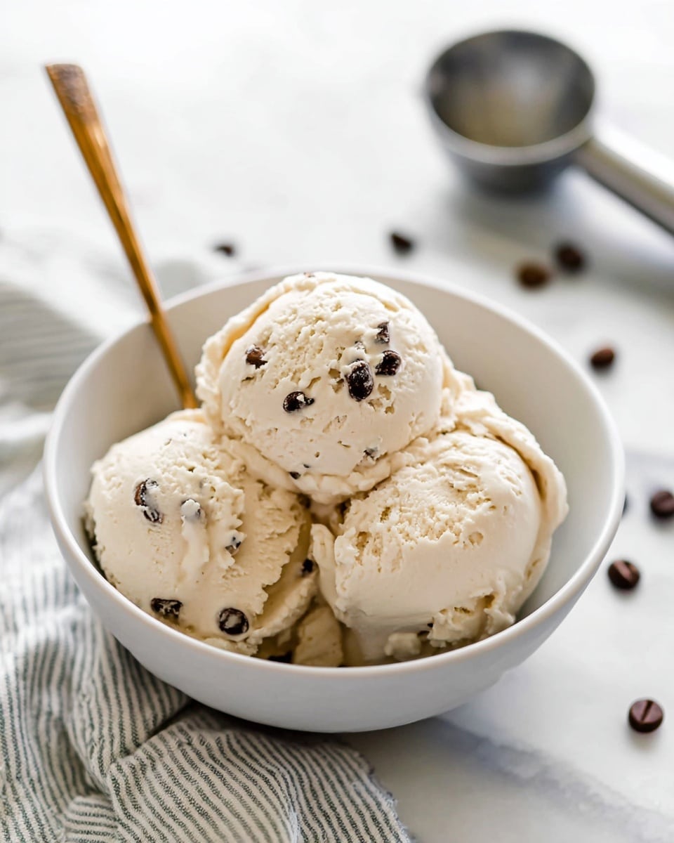 A white bowl filled with three scoops of creamy light beige ice cream that has small dark chocolate chips mixed in. The ice cream looks smooth with soft swirls around the edges of each scoop. A silver spoon with a golden handle rests inside the bowl, angled toward the front. The bowl sits on a white marbled surface with a light-colored striped cloth underneath, scattered with a few dark chocolate chips. In the blurred background, a metal ice cream scoop lies on the white marbled surface. Photo taken with an iphone --ar 4:5 --v 7