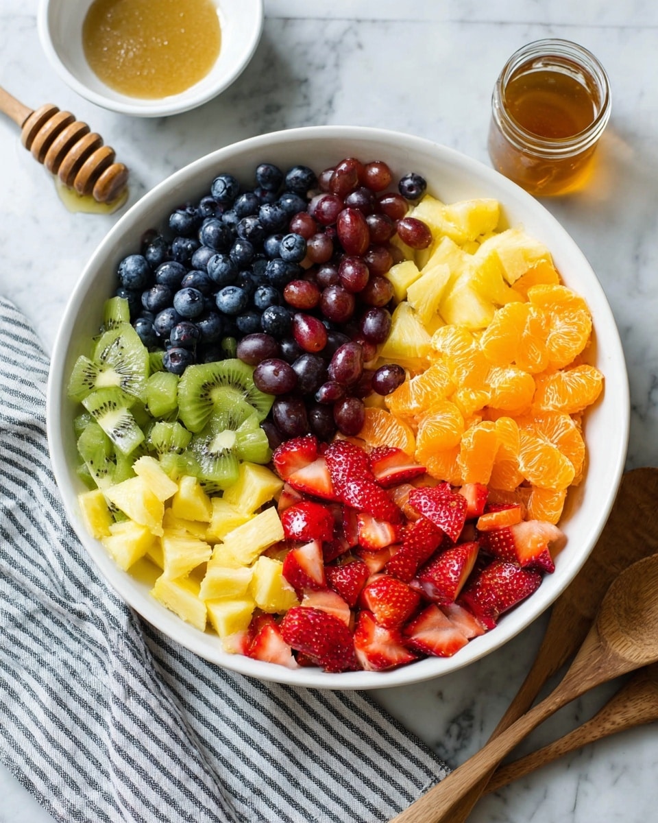 A white bowl holds six sections of fruit arranged in neat layers: deep blue blueberries form one layer at the top center, dark red grapes sliced in half create a shiny layer to the right, bright red strawberries cut into halves and quarters fill the bottom right, vibrant orange mandarin slices make a layer at the bottom center, light yellow pineapple chunks are placed on the bottom left, and fresh green kiwi pieces form the top left section. The bowl sits on a white marbled surface with a gray striped cloth nearby, wooden salad utensils resting to the right, a white bowl with a light brown dressing on the left, and a jar of honey with a honey dipper in the background. photo taken with an iphone --ar 4:5 --v 7