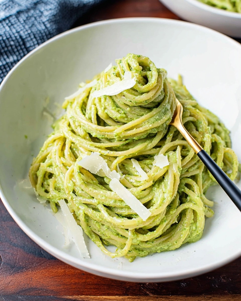 A white shallow bowl holds a mound of spaghetti thickly coated in creamy bright green pesto sauce. The pasta is twisted into a small heap in the center, showing off the sauce’s smooth texture mixed with small bits of basil and seasoning. Thin, pale shavings of parmesan cheese are scattered on top, adding a touch of contrast. A black and gold fork is stuck into the right side of the pasta, the fork partially wrapped with a few noodles. The bowl sits on a dark wooden surface with a hint of a blue checkered cloth in the upper background. photo taken with an iphone --ar 4:5 --v 7