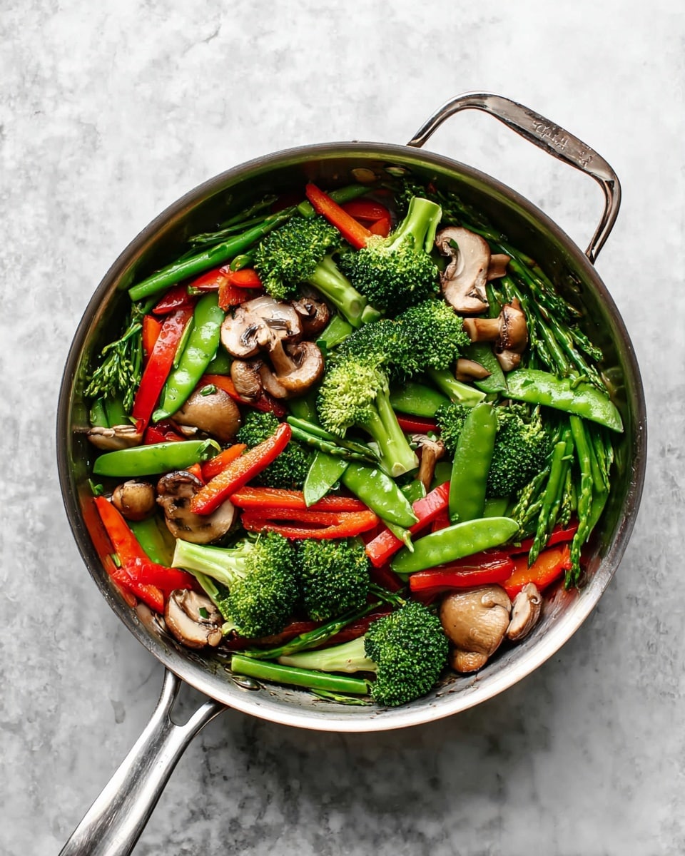 A shiny metal pan filled with a colorful mix of vegetables sits on a white marbled surface. The pan contains bright green broccoli florets and snap peas, long thin green asparagus pieces, red bell pepper strips, and brown mushroom halves scattered throughout. The vegetables appear fresh and lightly cooked, showing firm textures and vibrant colors, arranged evenly within the pan. The pan has two handles, one short and one long, with the short handle on the left and the long handle angled to the right. photo taken with an iphone --ar 4:5 --v 7