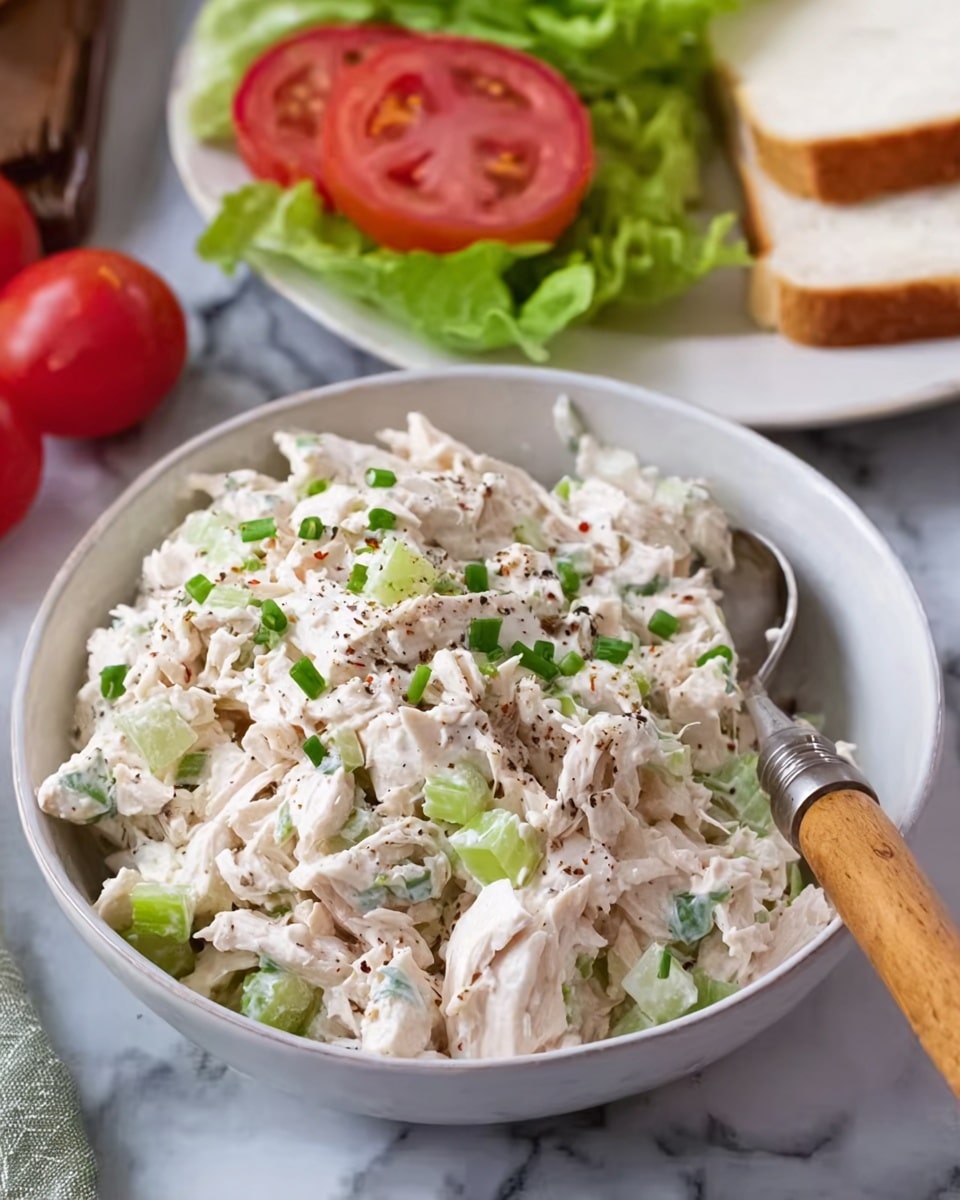 A white bowl filled with creamy chicken salad made of chopped white chicken pieces mixed with small green celery bits and sprinkled with black pepper and chopped chives, a silver spoon with a wooden handle rests inside the bowl. Behind, there is a white plate with green lettuce topped with bright red tomato slices and some white sandwich bread. The setting is on a white marbled surface with some halved red tomatoes visible in the background. photo taken with an iphone --ar 4:5 --v 7