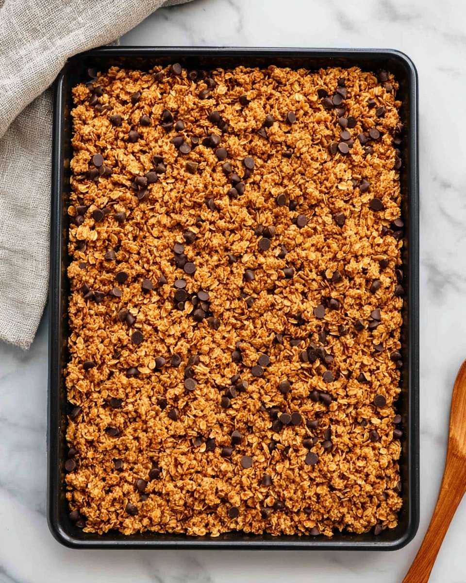 A rectangular black baking tray filled with a large, flat layer of golden brown granola mixed with small dark chocolate chips scattered throughout the surface. The granola has a rough and crumbly texture, with a mix of oats and small clustered bits, evenly spread to cover the entire tray. The tray is placed on a white marbled surface with a light gray cloth in the upper left corner and a wooden spoon to the right. The image shows a top-down view of the granola bars photo taken with an iphone --ar 4:5 --v 7