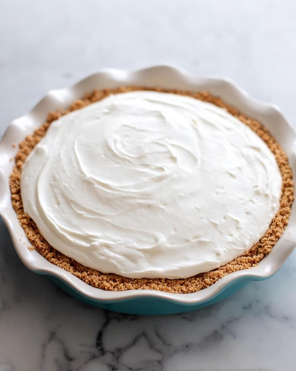 A round two-layer pie sits in a white pie dish with a slightly wavy edge. The bottom layer is a crumbly, light brown graham cracker crust with a coarse texture. The top layer is a thick, smooth white cream spread evenly and softly swirling across the surface. The pie dish is on a white marbled surface. The lighting is soft, showing the pie close up from the side. Photo taken with an iphone --ar 4:5 --v 7
