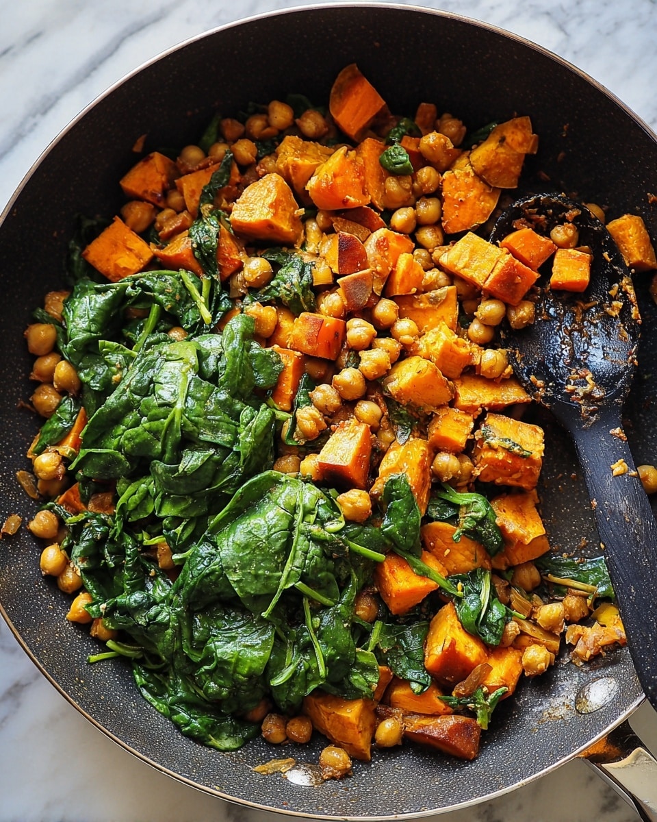 A close-up view of a dark non-stick pan filled with a colorful mix of cooked food, showing three main layers: the bottom layer has golden chickpeas, the middle layer consists of bright orange sweet potato cubes with a slightly crispy texture, and the top layer is fresh, dark green spinach leaves that look soft and wilted. The ingredients are mixed together evenly, showing their natural colors and textures. A black spoon covered in bits of the dish rests on the right side inside the pan, on a white marbled surface. photo taken with an iphone --ar 4:5 --v 7