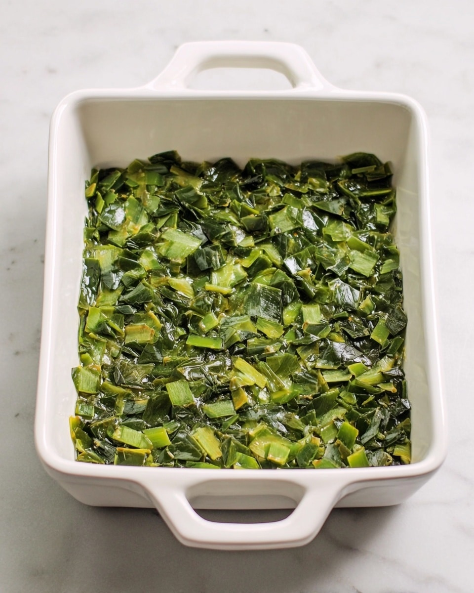 A white square baking dish with two handles is shown from above, resting on a white marbled surface. Inside the dish is a single even layer of small chopped pieces of green vegetables that appear soft and cooked, with varying shades of green from dark to light, creating a slightly shiny and moist texture. The pieces are spread out evenly across the bottom of the dish, covering the entire base without overlapping too much. Photo taken with an iphone --ar 4:5 --v 7