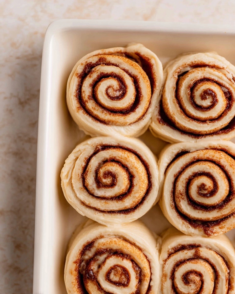 The image shows six unbaked cinnamon rolls arranged in a close group on a white rectangular pan. Each roll has a light beige dough with dark brown cinnamon sugar spirals forming circular layers from the center to the edge. The dough looks soft and slightly shiny, indicating it is ready to be baked. The white pan sits on a white marbled surface with soft, light gray veining. The cinnamon rolls are placed close enough to slightly touch each other, filling the pan. Photo taken with an iphone --ar 4:5 --v 7
