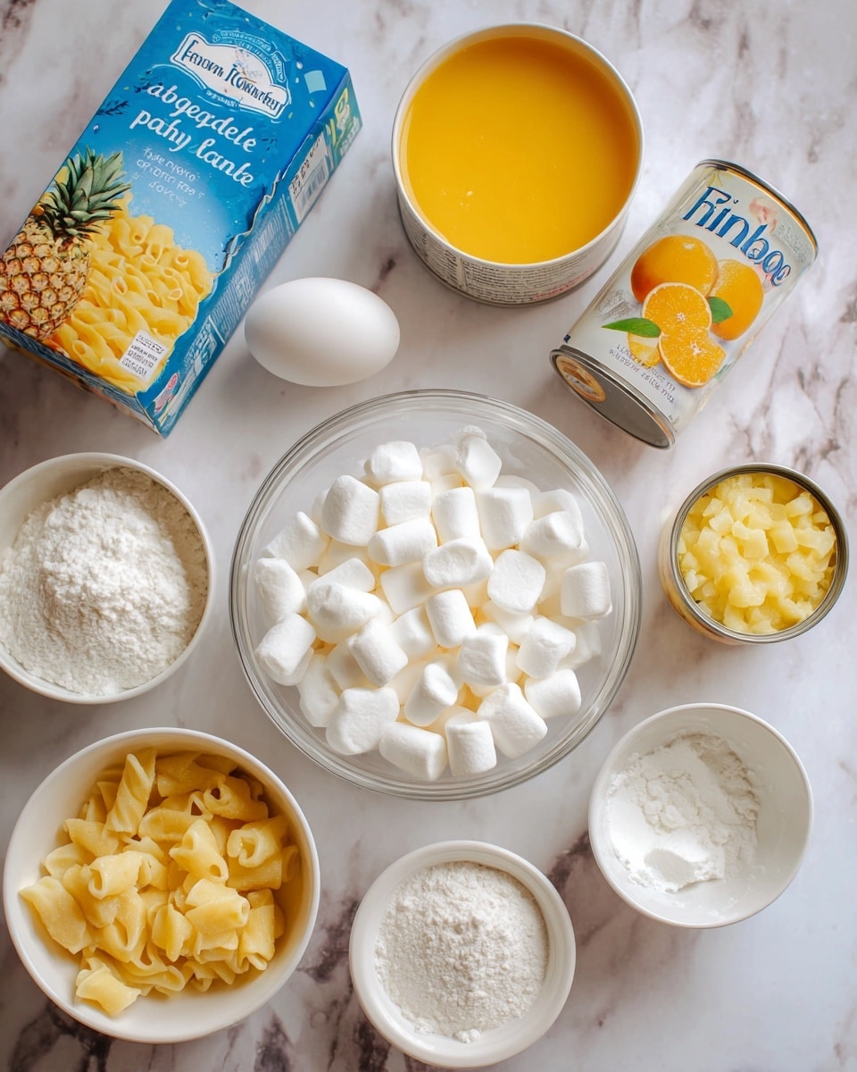 The image shows various ingredients arranged on a white marbled surface, including a clear bowl filled with many small white marshmallows at the center. Near the marshmallows are an unopened white egg, a white bowl with smooth white cream, and a small white bowl with a white powder, likely baking powder. To the left, there is a blue and yellow box of pasta, beside an open can filled with yellow pineapple chunks. At the top, there is a white bowl containing yellow-orange juice or liquid and two open cans, one with orange mandarin slices and the other with crushed light yellow pineapple. A small white bowl filled with white sugar is also visible. The text
