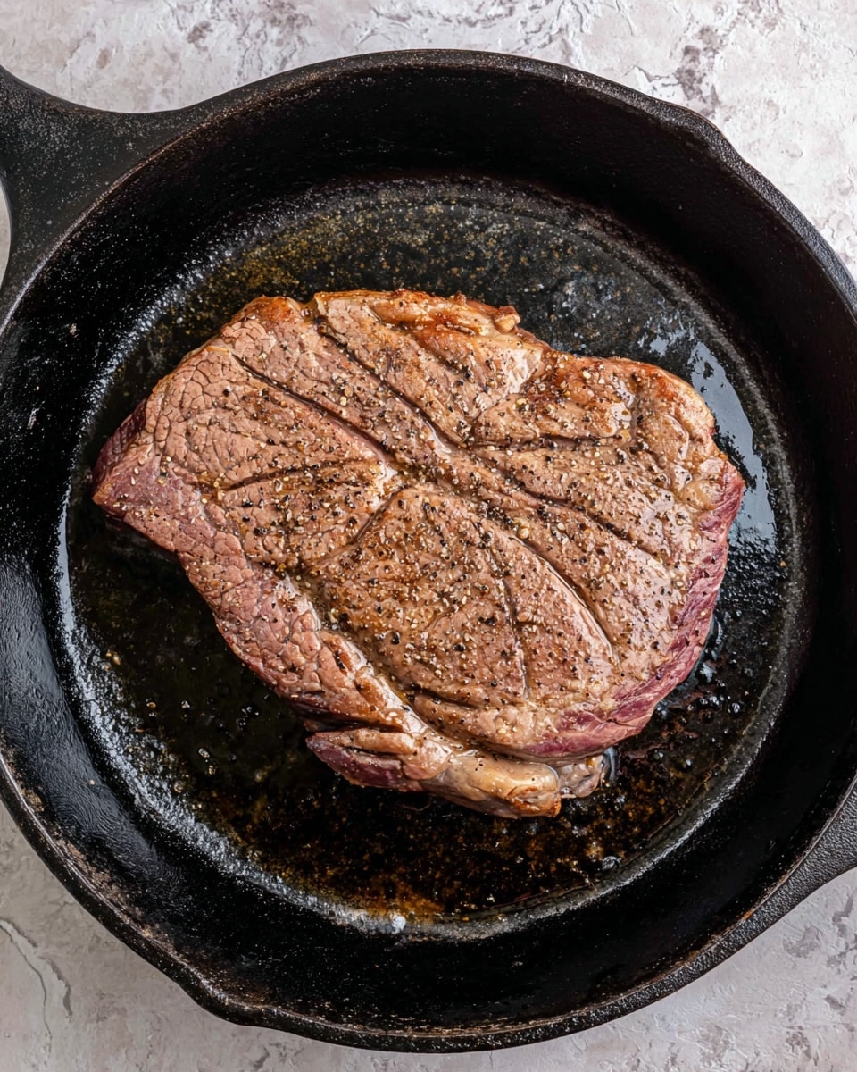 A single thick steak with a brown and slightly pink color is cooking in a black cast iron pan. The steak has a textured surface with specks of black pepper and some seared lines, showing a juicy and tender look with some fat on the edges. The cast iron pan is black and shiny with cooking oil visible around the steak. The pan sits on a surface with a white marbled texture photo taken with an iphone --ar 4:5 --v 7