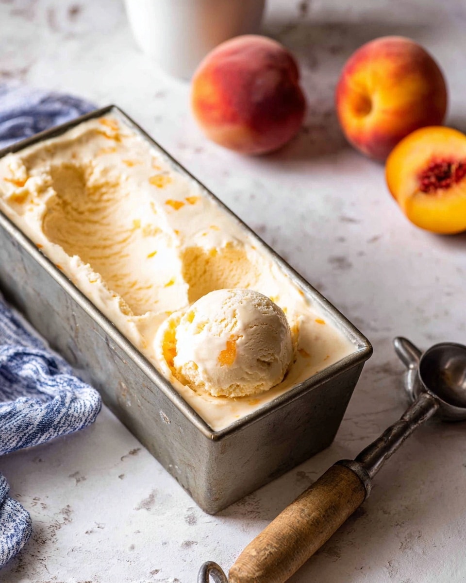 A rectangular metal container filled with smooth, creamy pale peach-colored ice cream with small orange specks, one scoop taken out showing soft texture inside; three whole peaches with reddish-yellow skin sit blurred in the background on a white marbled surface, next to a white cup; an old-fashioned metal ice cream scooper with a wooden handle lays in the foreground, the scene has cozy soft lighting, photo taken with an iphone --ar 4:5 --v 7
